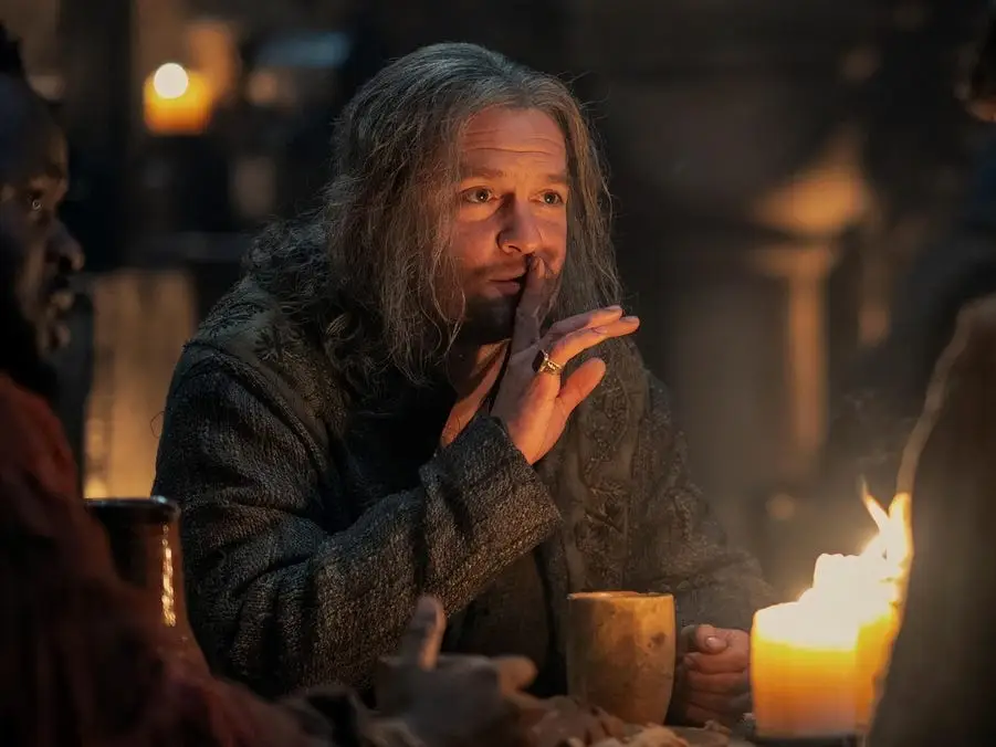 tom bennett as ulf the white in house of the dragon. he's a middle aged man with shoulder length grey hair, placing a hand up to his lips as he sits at a tavern table with a group of men