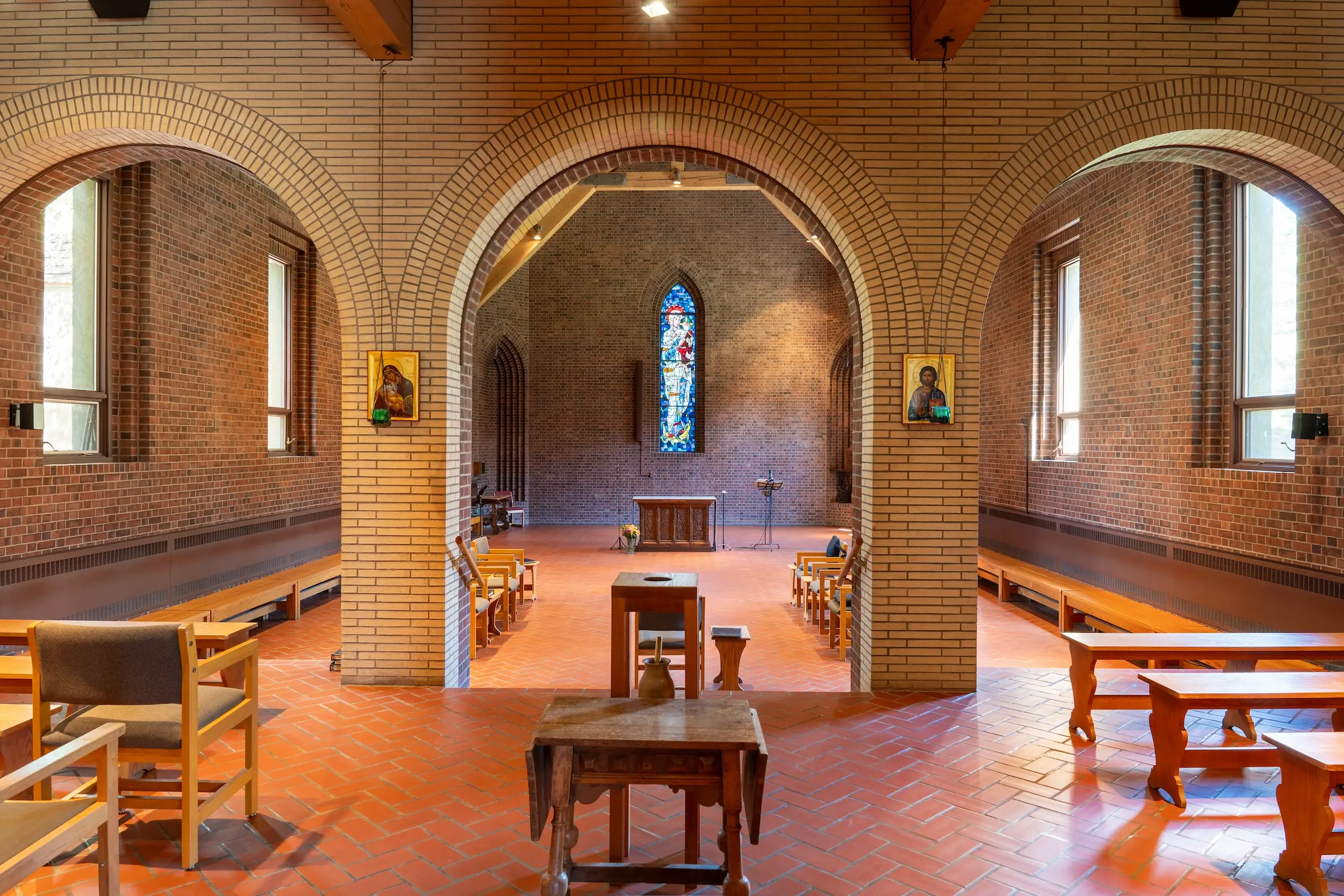 The interior of a room with wooden benches, wooden chairs, an altar, two portraits of religious icons, and a stained-glass window.