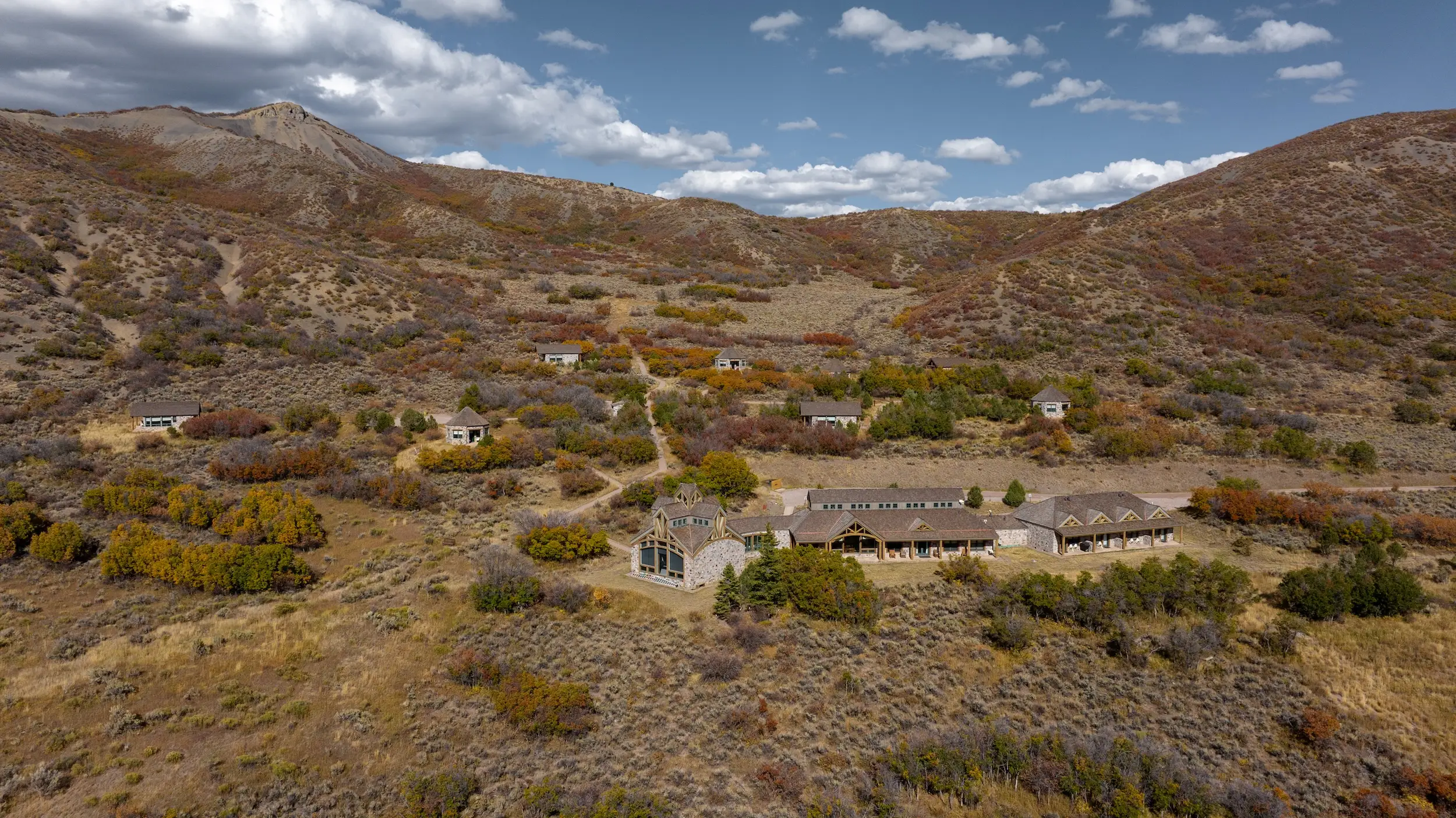 An aerial shot of some buildings on a spacious piece of land between mountains, with dry ground and brown vegetation.