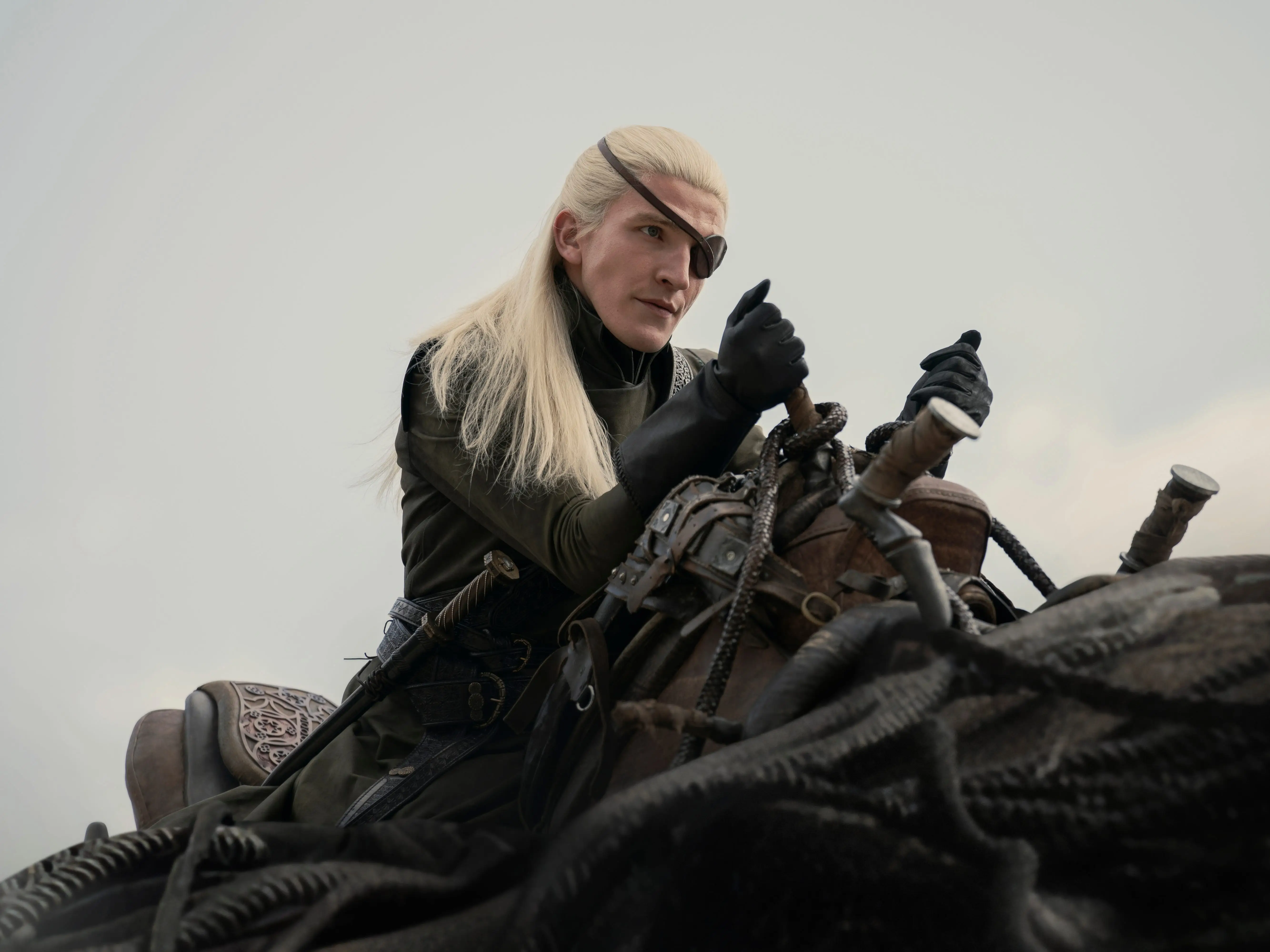 ewan mitchell as aemond targaryen, mounted on top of a dragon and holding on to a saddle. his silver hair is pulled half back and he's wearing an eye patch, clad in all black as he holds onto the saddle handles with gloved hands.