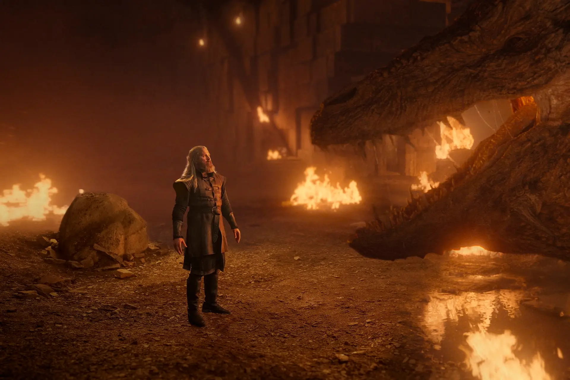 kieran bew as hugh hammer in house of the dragon. he's a middle aged man with long blonde hair wearing dark robes, standing in front of the open maw of vermithor, a very large dragon. around him, there are various fires alight