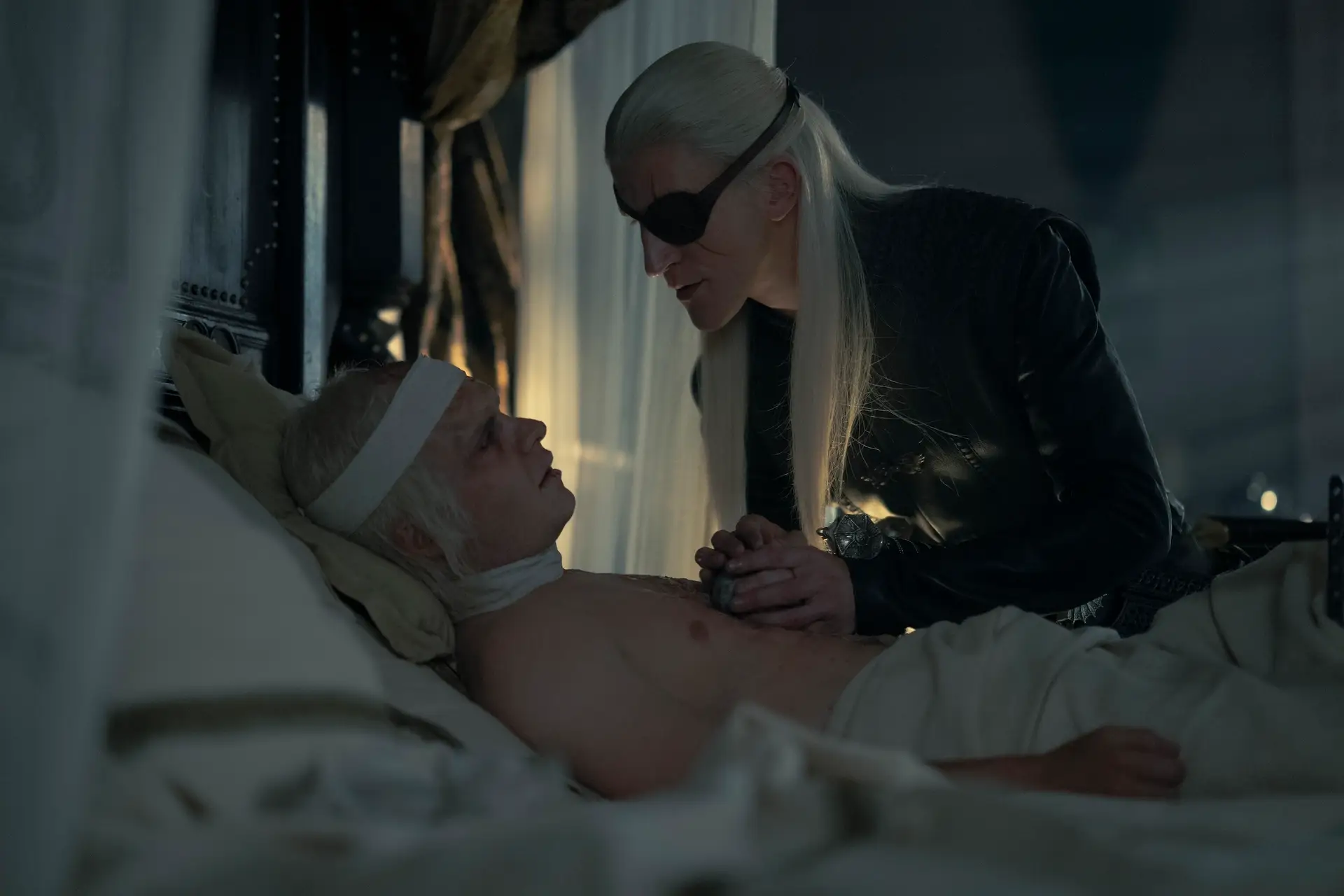 tom glynn-carney as aegon in house of the dragon, laying shirtless and bandaged, his blonde hair stringy and thin, in a lavish bed. his brother aemond, played by ewan mitchell, is standing above him, placing a stone ball into aegon's hand and looming over his hcest. he wears all black, a black eyepatch, and has long blonde hair