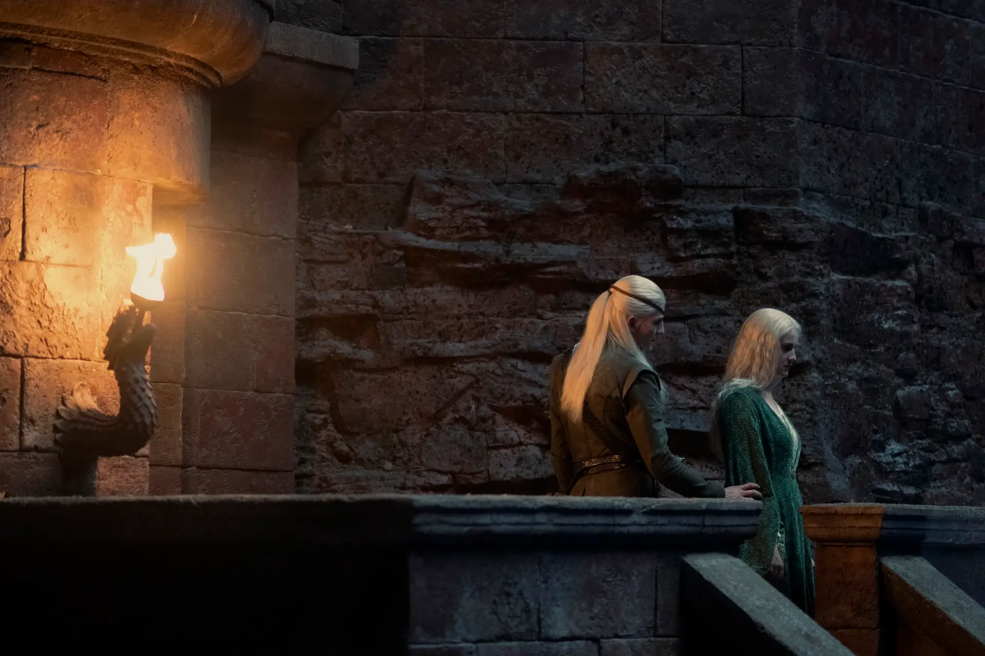ewan mitchell and phia saban as aemond and helaena targaryen, standing on a stone balcony and illuminated by a torch. aemond is wearing black clothes, while helaena is in a green dress. both have long blonde hair