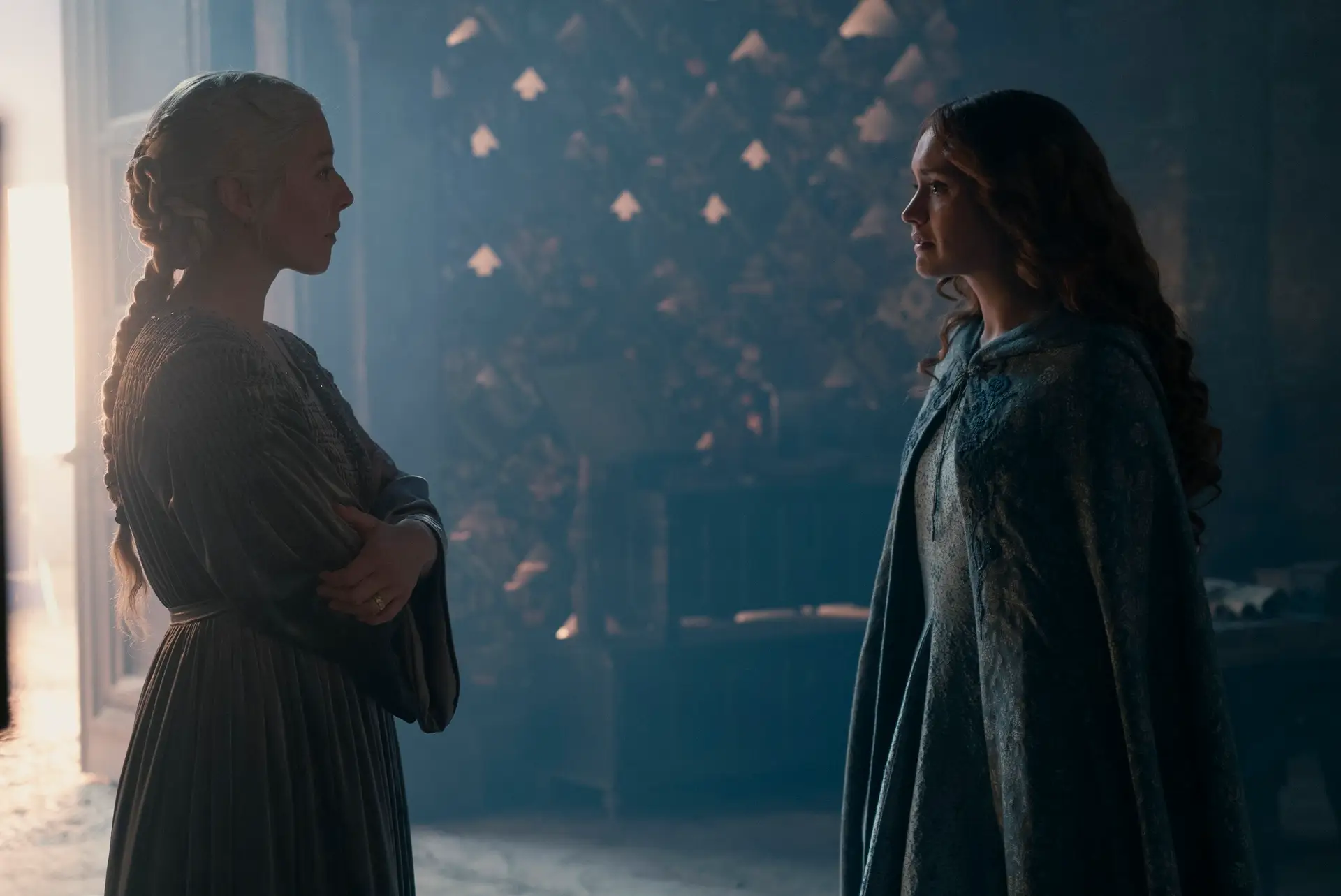 emma d'arcy and olivia cooke as rhaenyra and alicent on house of the dragon. rhaenyra is wearing a flowing robe, her hair pulled back in a braid, while facing alicent, clad in a blue dress and traveling cloak. the two are looking emotionally towards each other