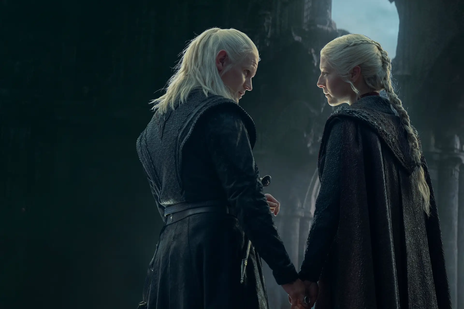 matt smith and emma d'arcy as daemon and rhaenyra targaryen. they're seen from the back, both of their faces turned toward each other, holding hands and starting into each others' eyes. both have blonde hair, daemon's worn shoulder length and half back, rhaenyra's long and in a braid