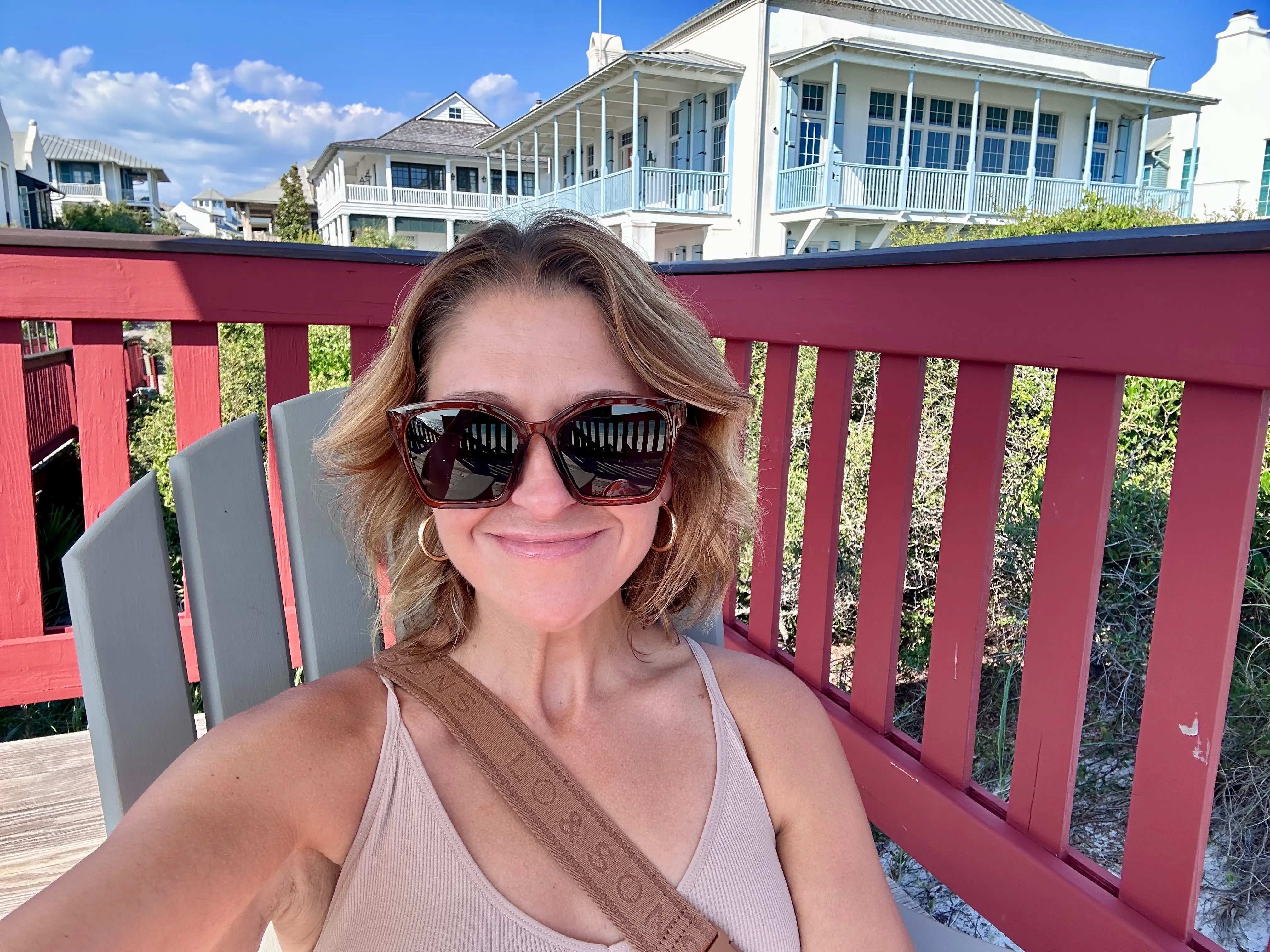 The author smiling and wearing sunglasses, taking a selfie as she sits on a chair outside.