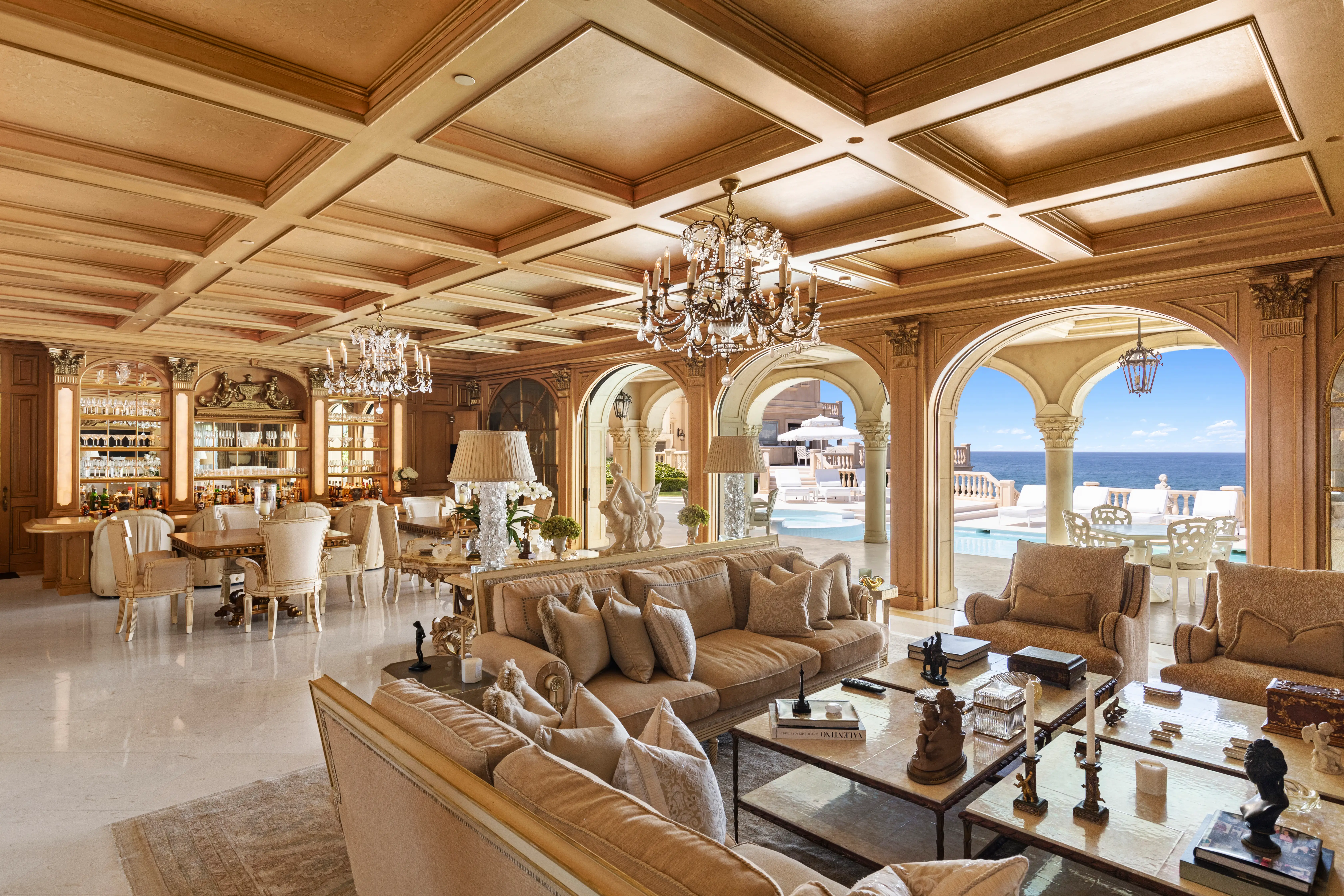 The living room of the Sand Castle mansion, featuring two crystal chandeliers, a dining and seating area, and views of the outdoor pool.