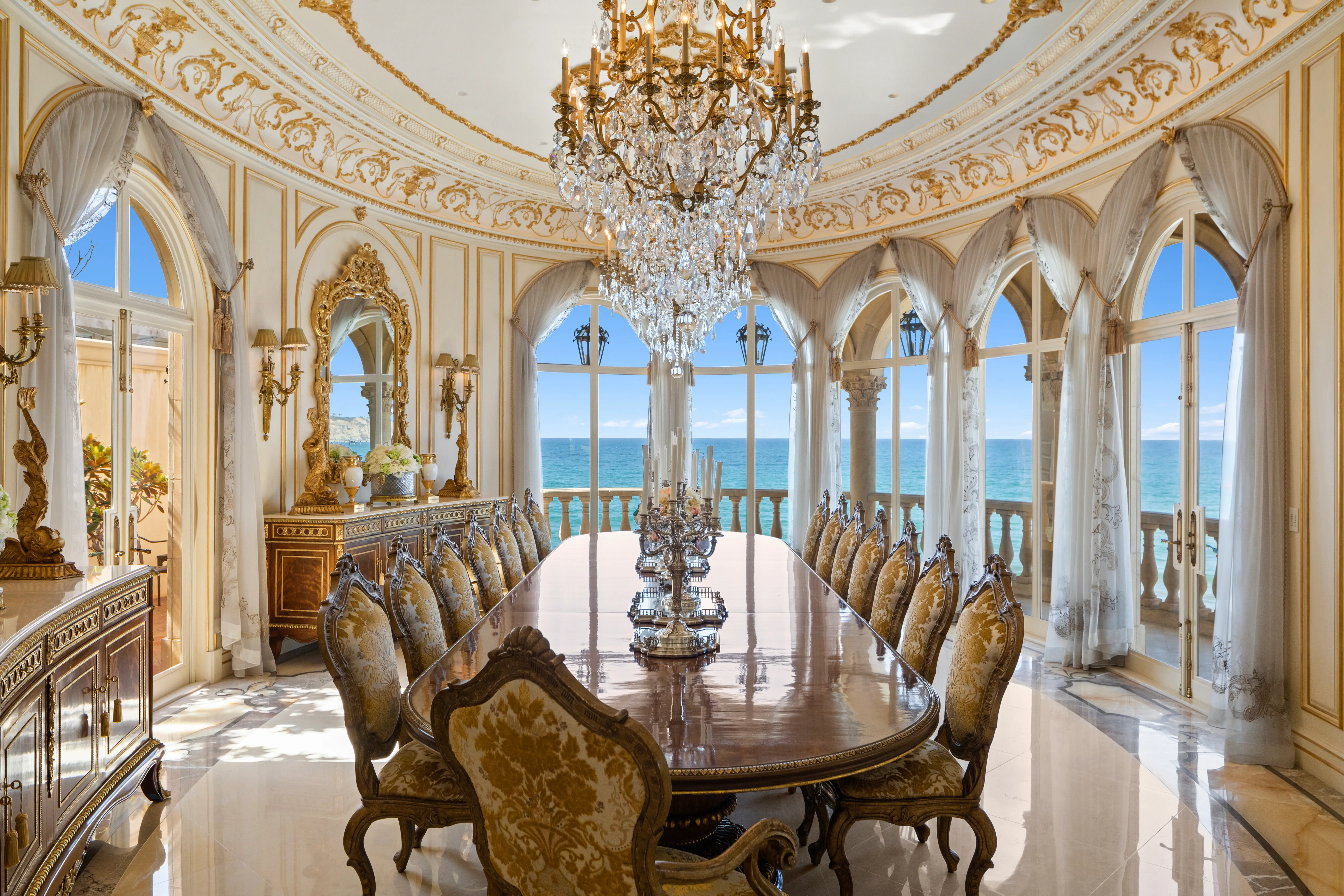 A long dining-room in the Sand Castle mansion underneath an elaborate crystal chandelier with gold accents, with the windows showcasing views of the ocean.