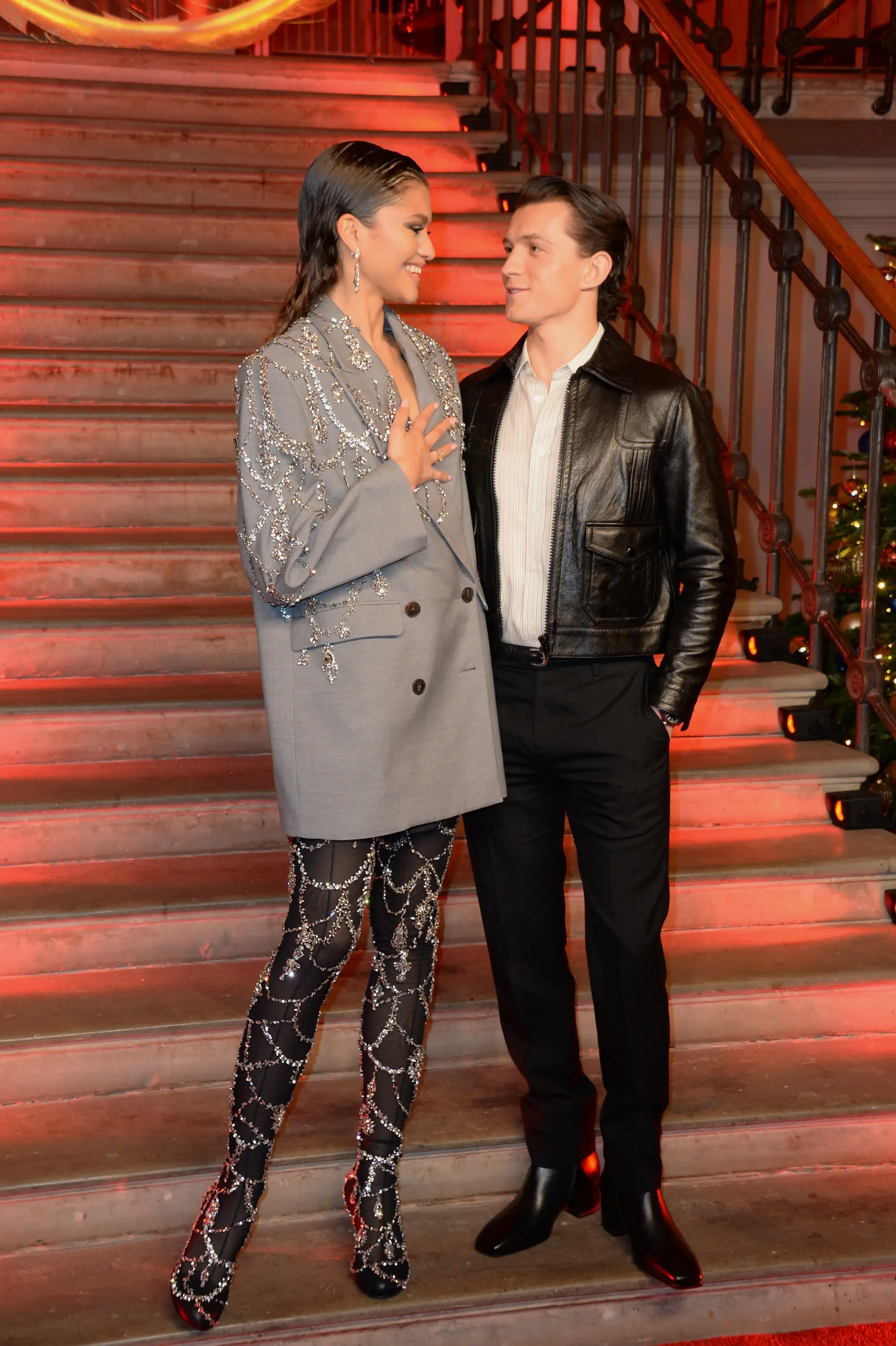 Zendaya and Tom Holland smile at each other as they stand on a staircase.