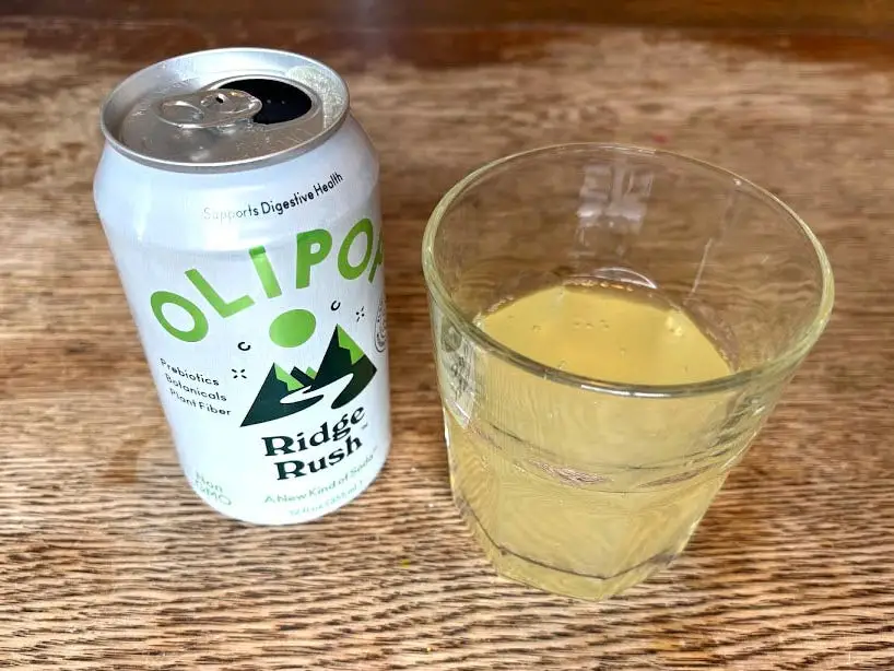 An open can of Ridge Rush Olipop next to a small, clear glass with yellow liquid inside. Both are sitting on a wooden table.