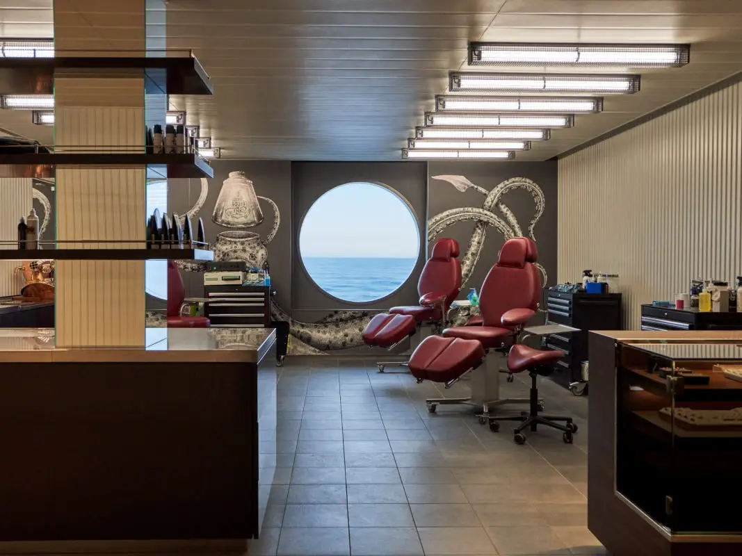 A tattoo parlor with red chairs and a port hole window with a view of the ocean on the right and a counter top and shelf on the left