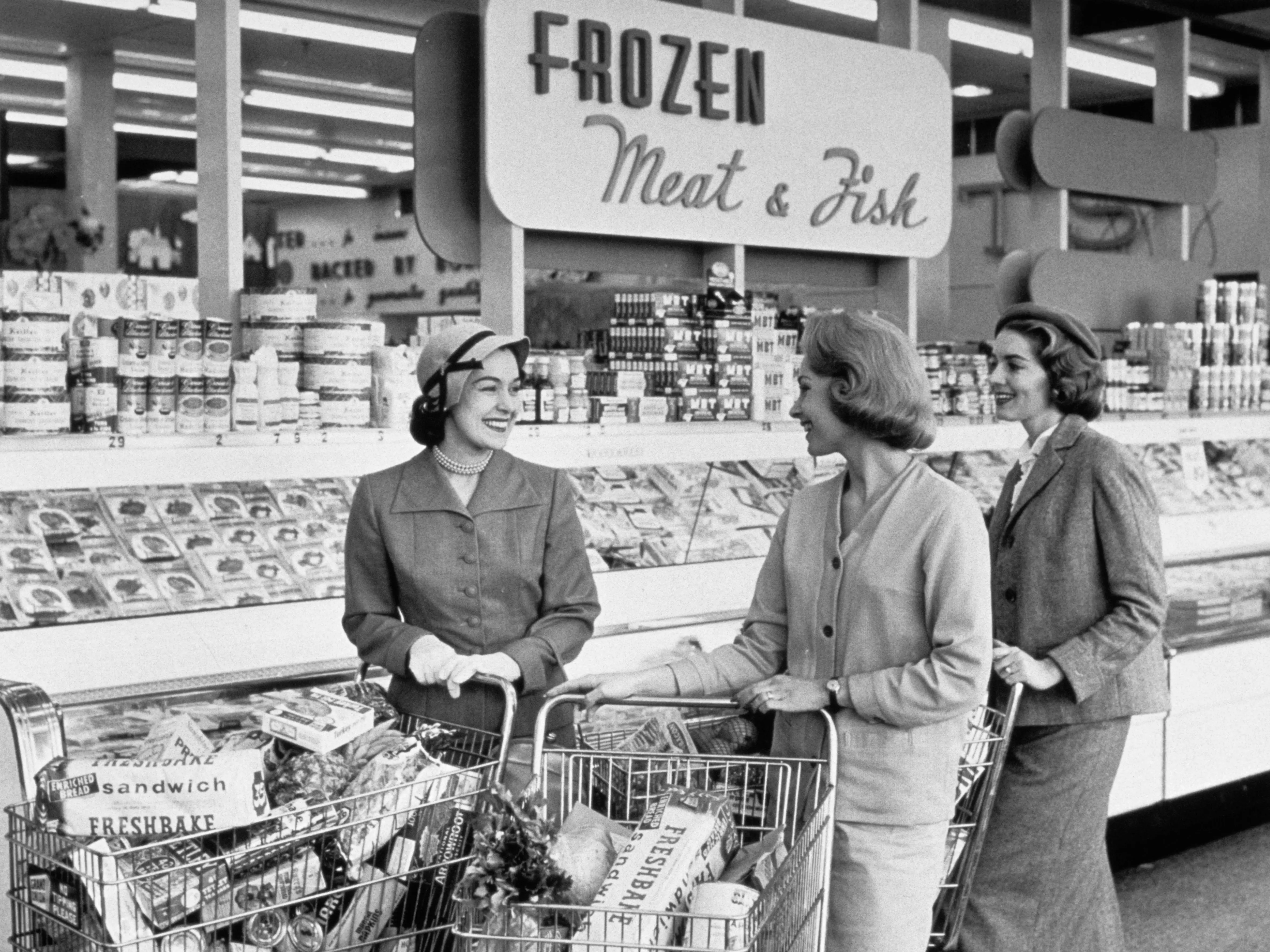 Three women push shopping carts in front of a case and a sing reading frozen meat and fish