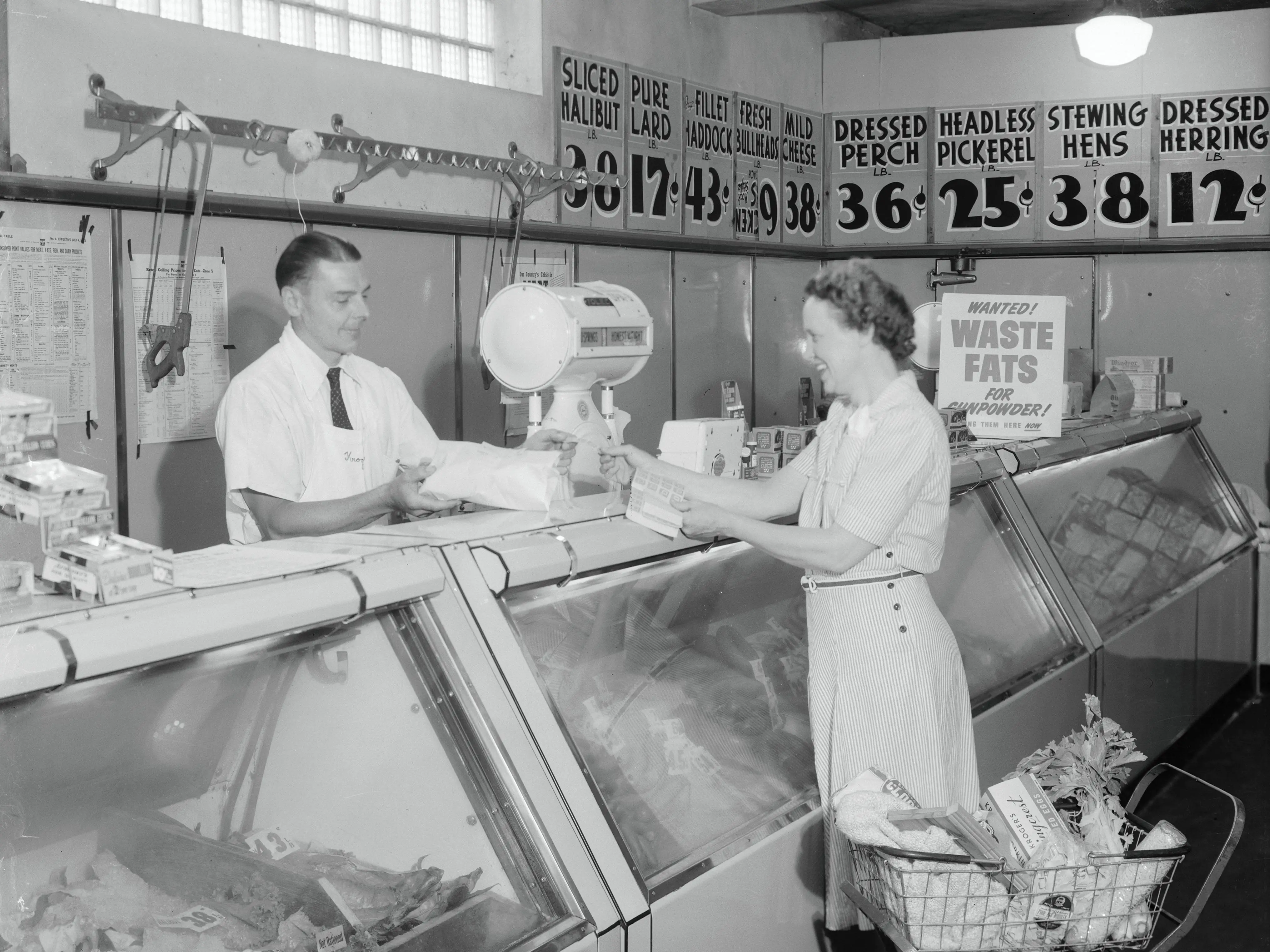 A woman stands next to a shopping cart while a butcher helps her at the meat counter