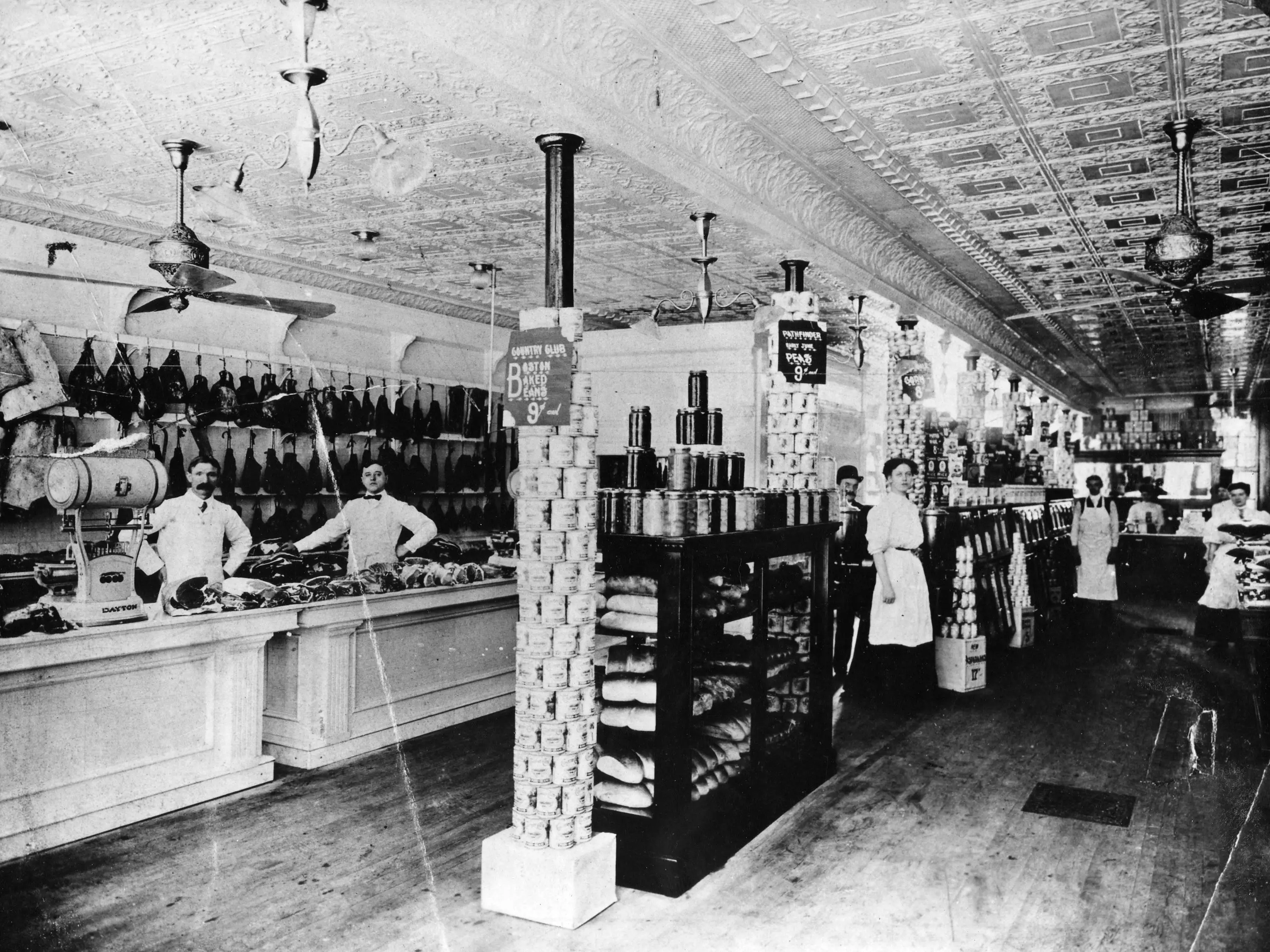 Several people stand in a general store with goods stacked up and on shelves