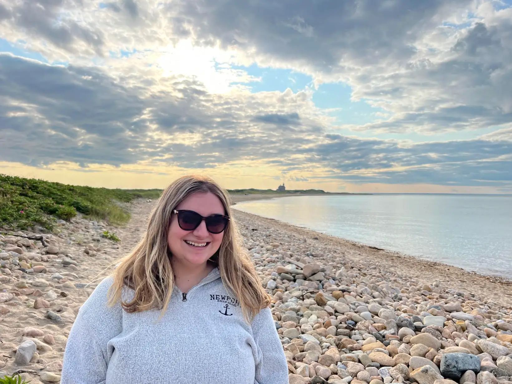 A woman standing on a pebble beach, smiling.