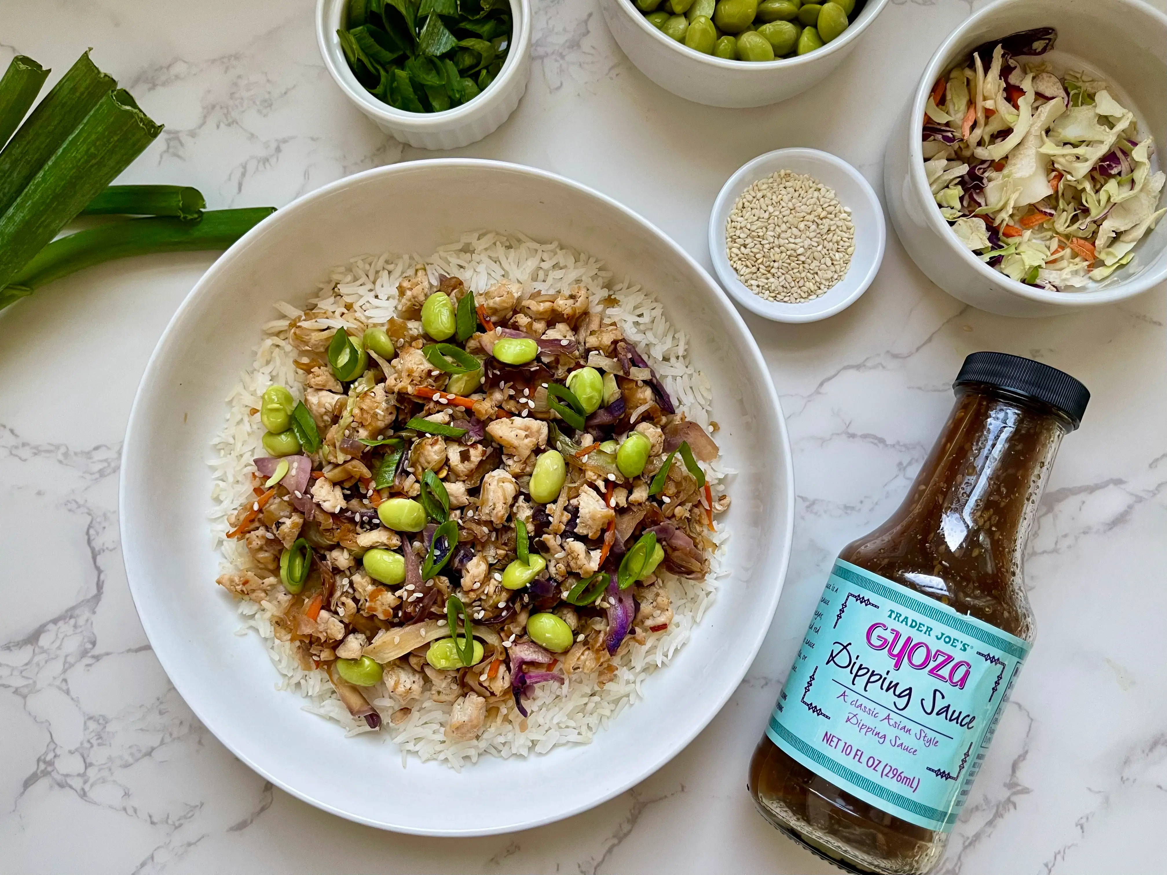 A rice bowl with turkey, edamame, and coleslaw on a table with bowls of ingredients and a bottle of Trader Joe's gyoza dipping sauce.