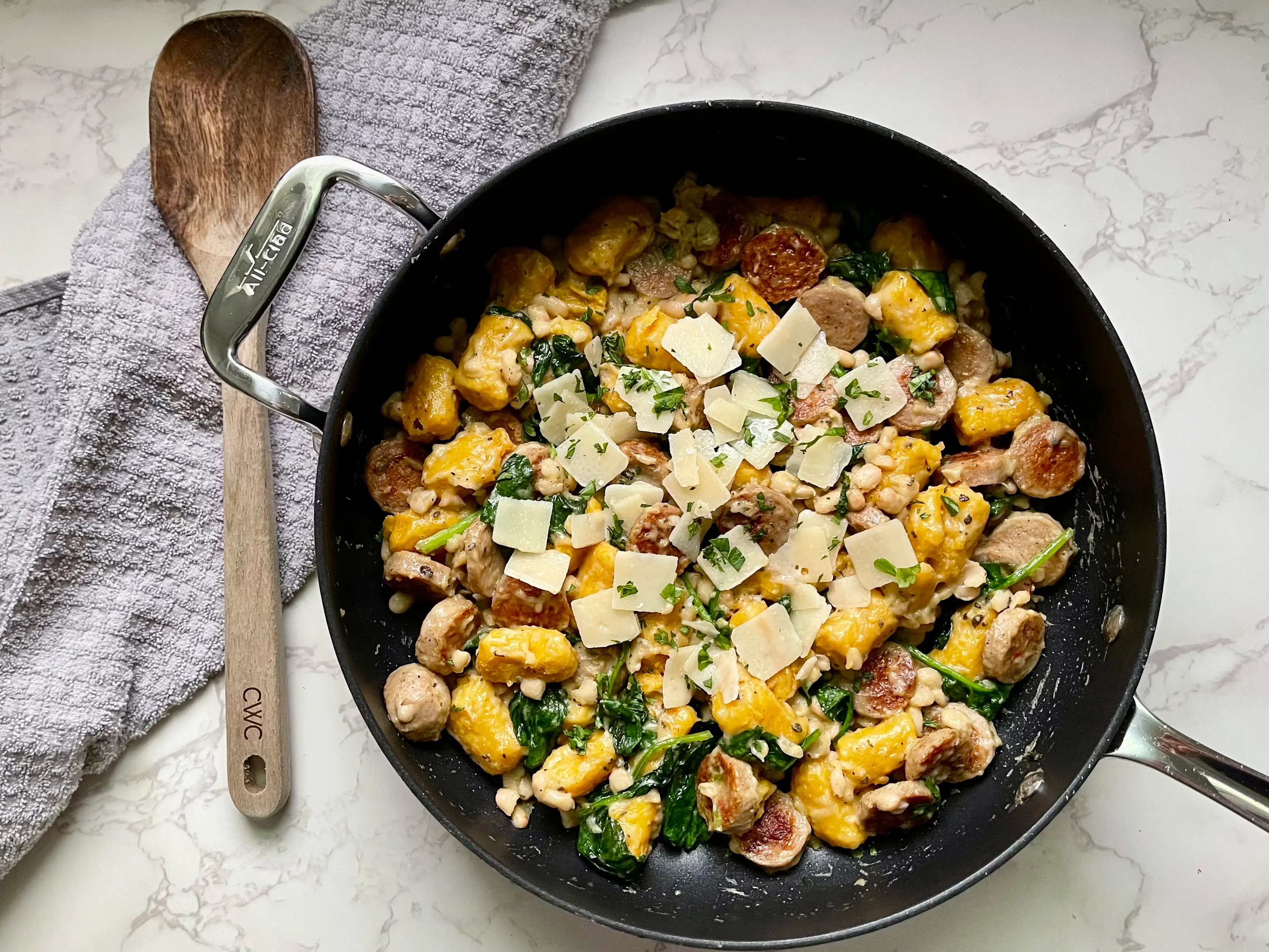 A skillet of sweet-potato gnocchi with spinach, sausage, and cheese, on a countertop next to a dish towel and a wooden spoon.