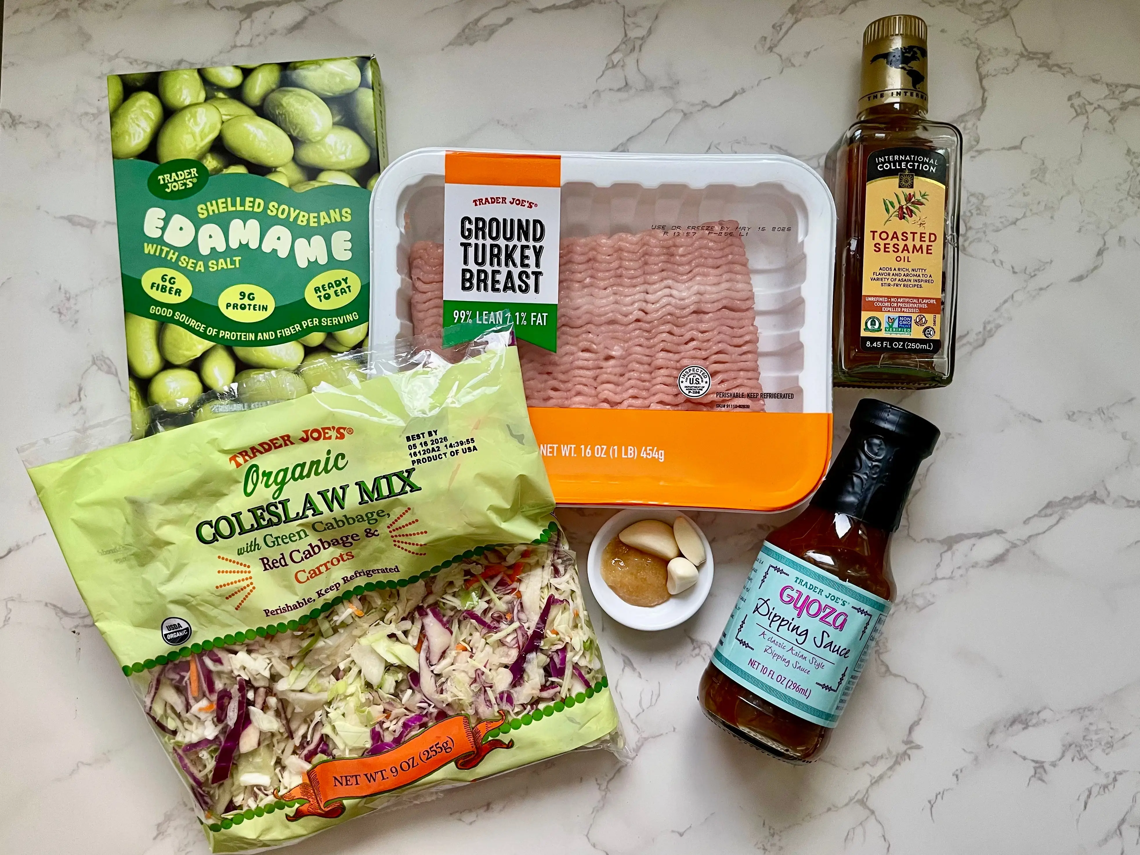 A box of edamame, a bag of coleslaw, a package of ground turkey,  a ramekin of garlic, a bottle of sesame oil, and a bottle of gyoza dipping sauce — all from Trader Joe's — on a countertop.