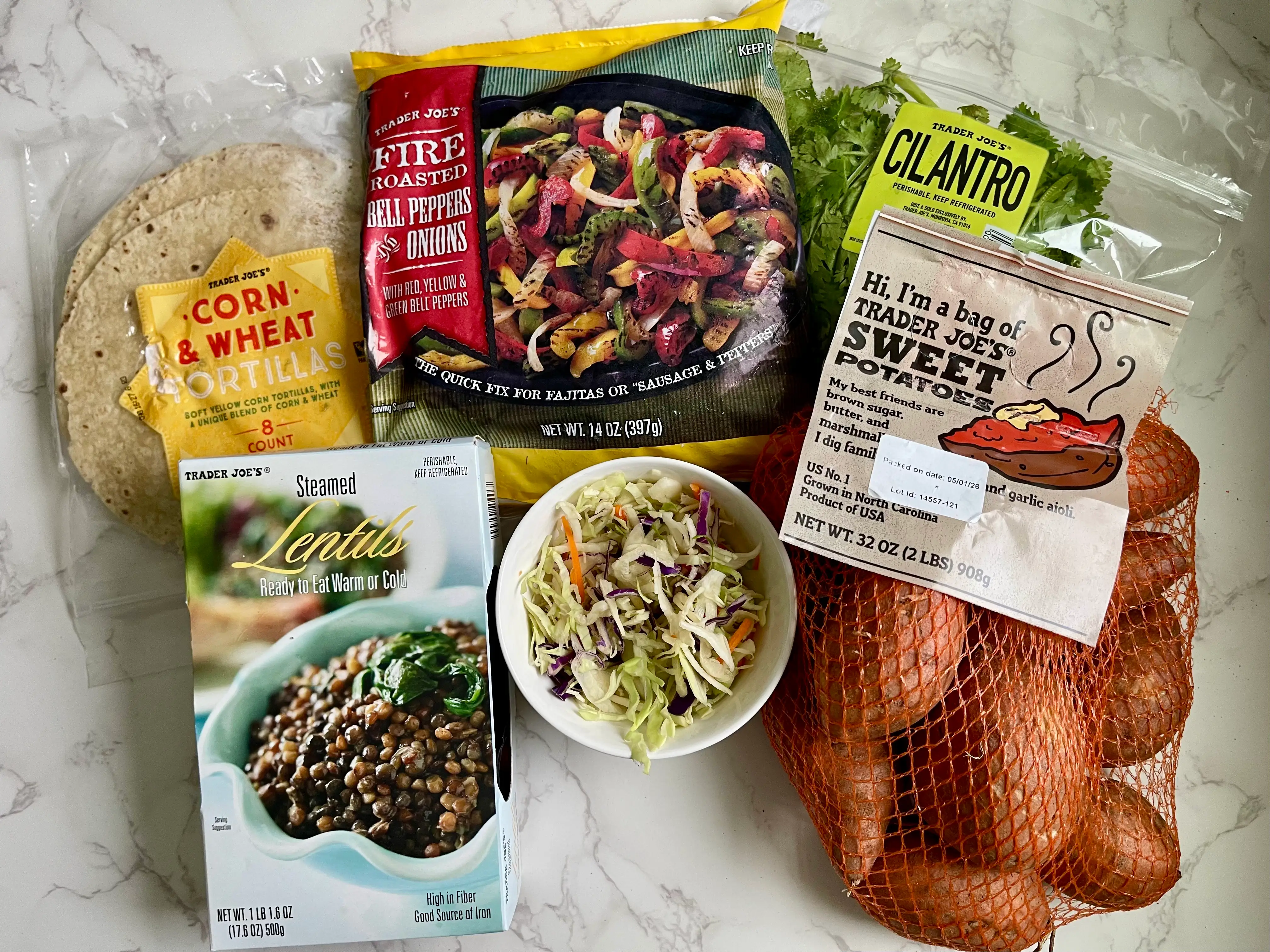 A package of corn tortillas, a bag of frozen veggies, a bowl of coleslaw, a package of cilantro, and a bag of sweet potatoes— all from Trader Joe's — on a countertop.