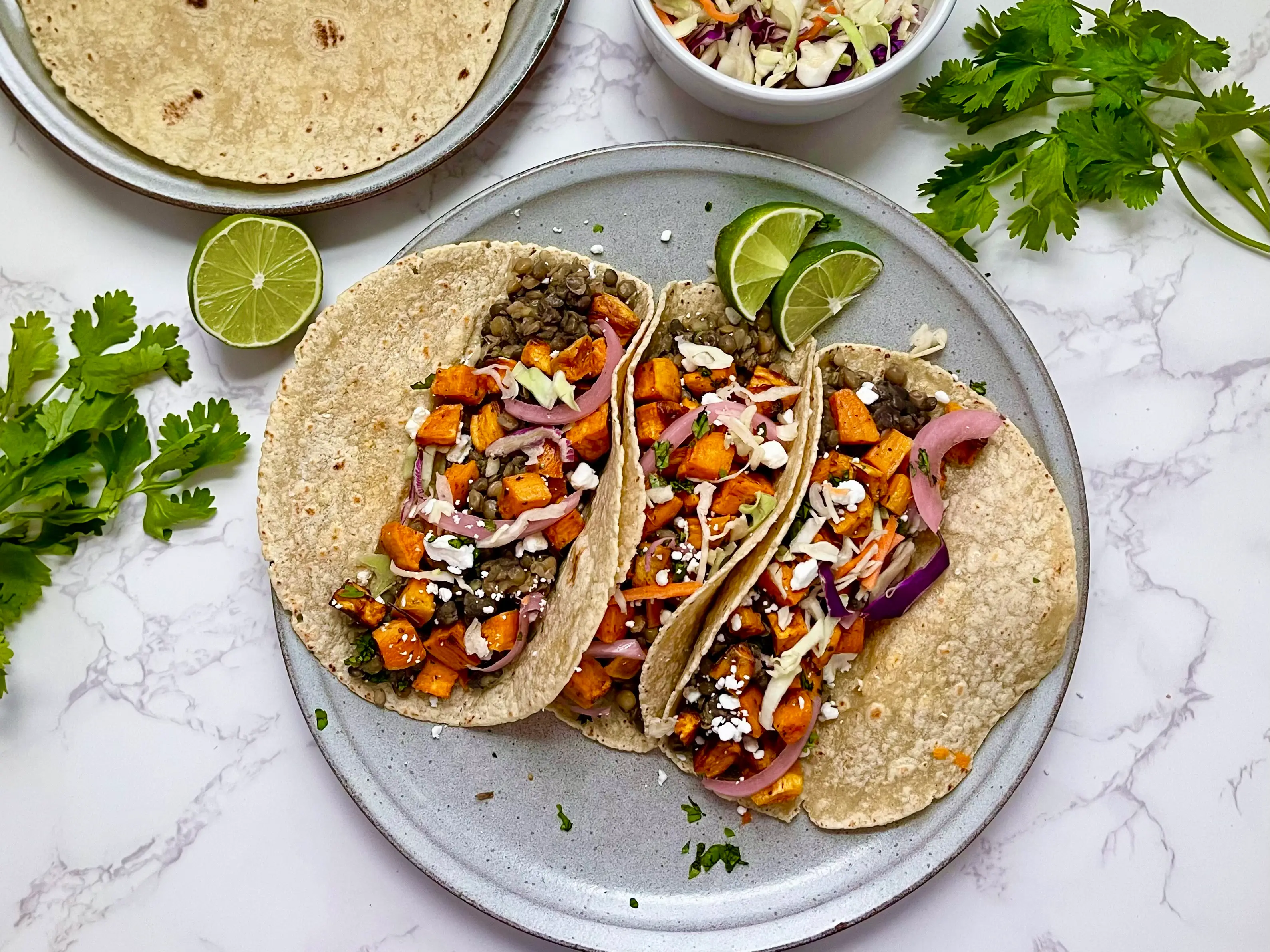 Three tacos, filled with sweet potatoes, lentils, onion, and cheese, on a plate.