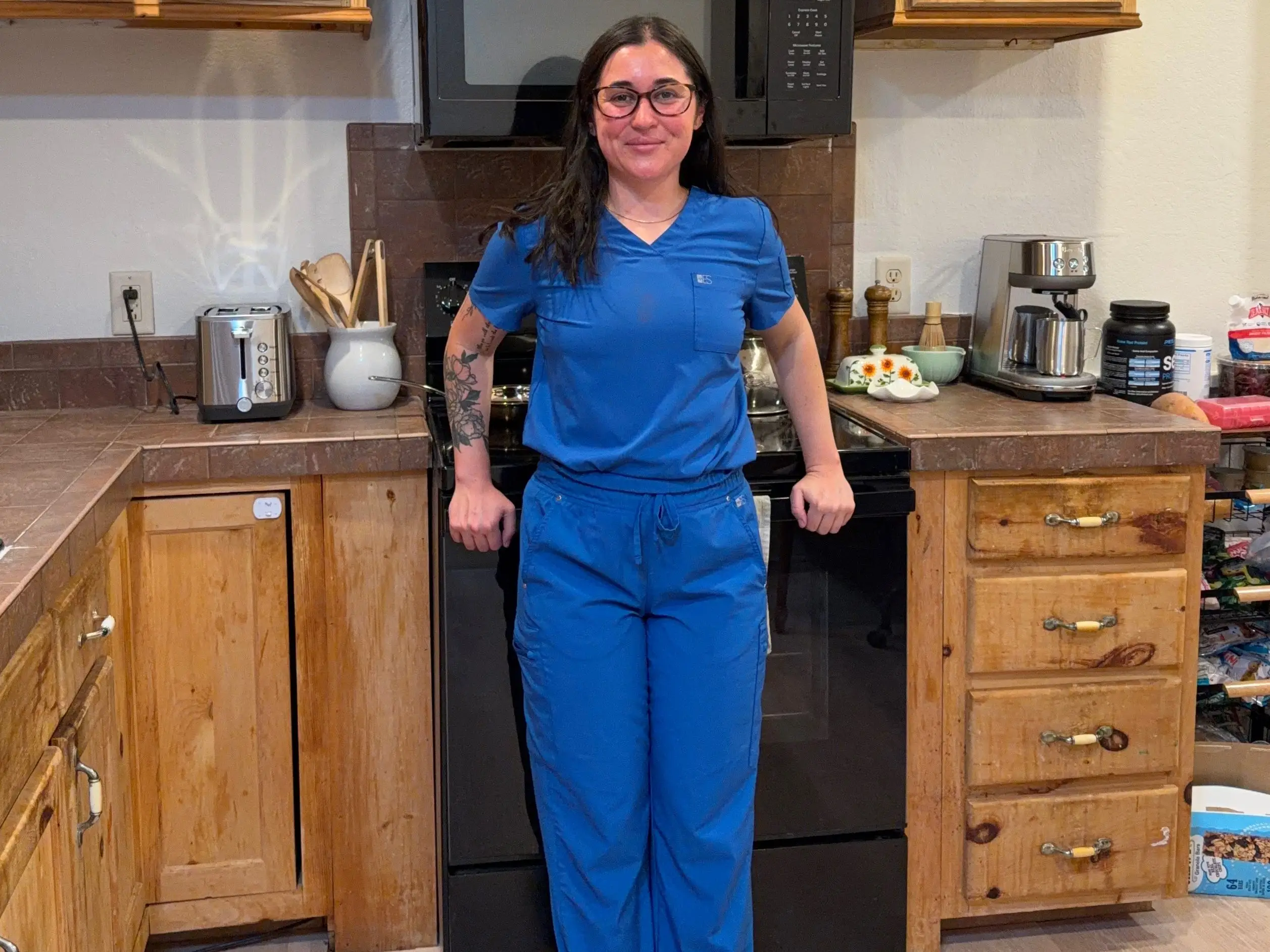A woman stands in a kitchen wearing blue scrubs.