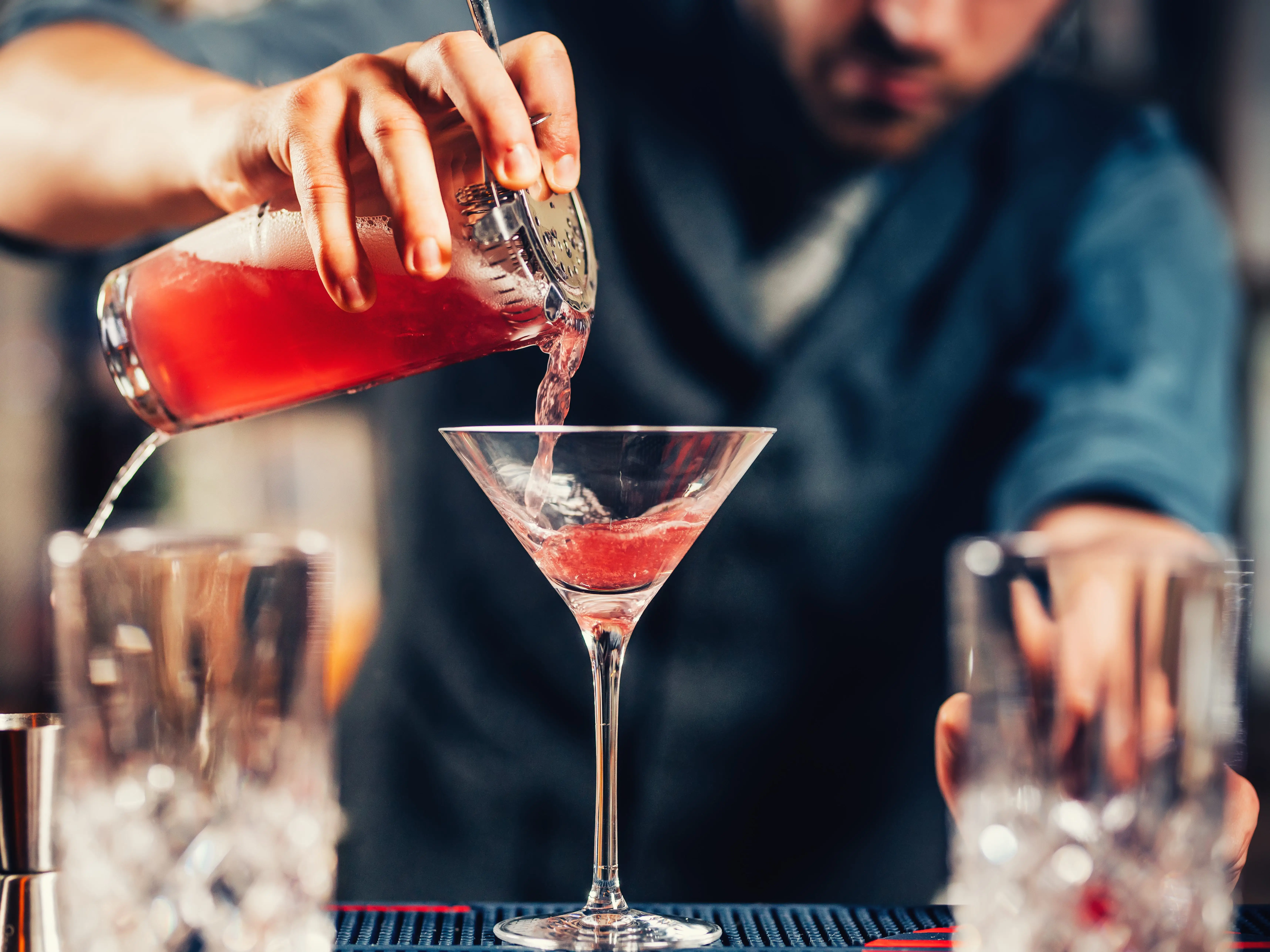 A bartender pours a pink cocktail from a shaker into a martini glass.