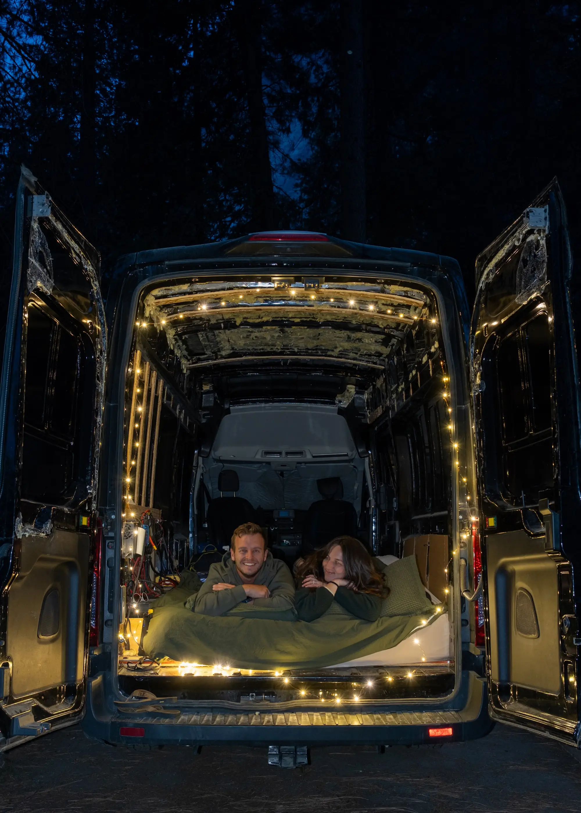 A couple lies down in a bed inside a van with lights in it.