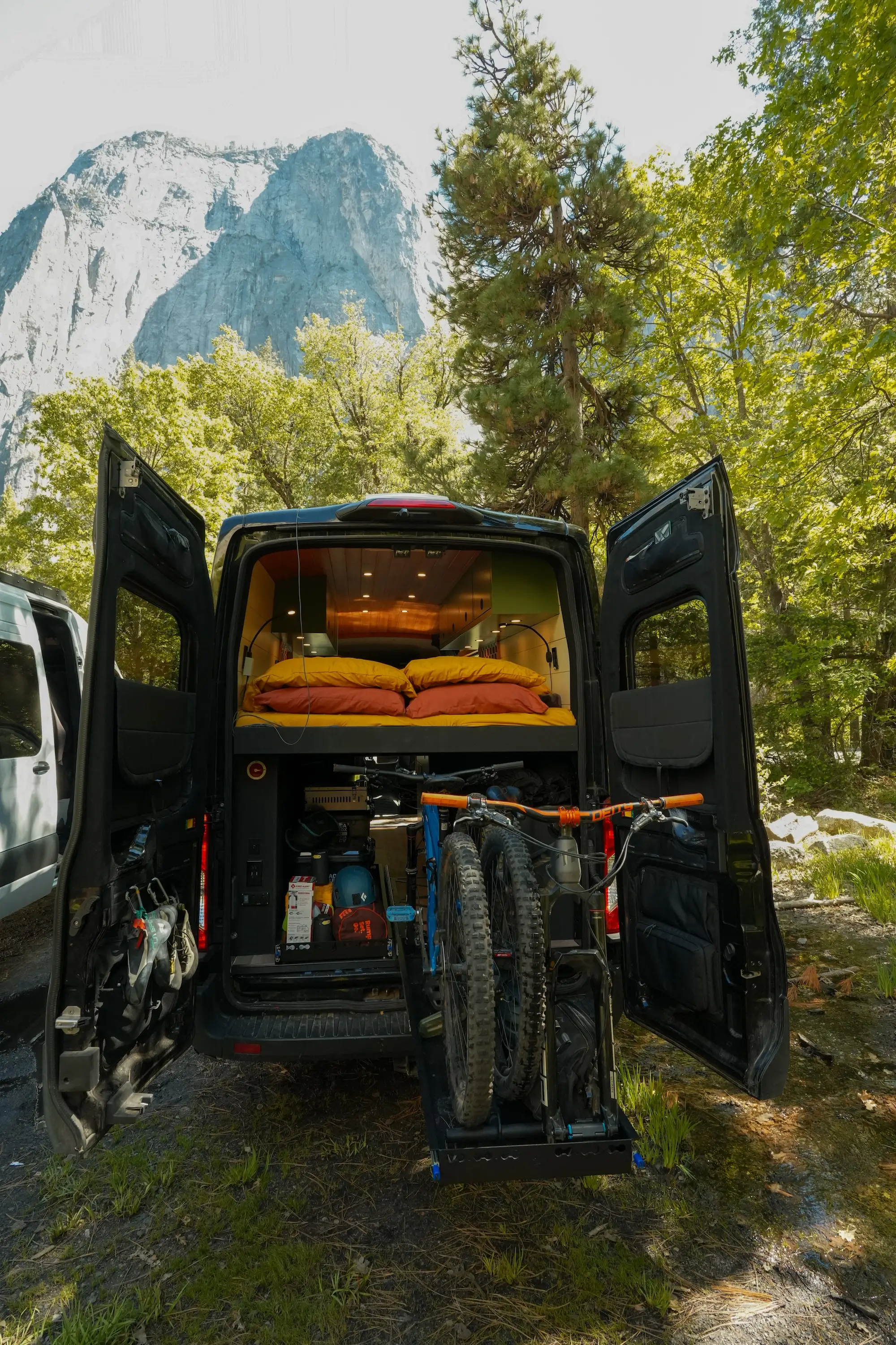The back of a van with the doors open. A bed sits atop the back, while a storage space sits under it with a mountain bike sliding out of it.