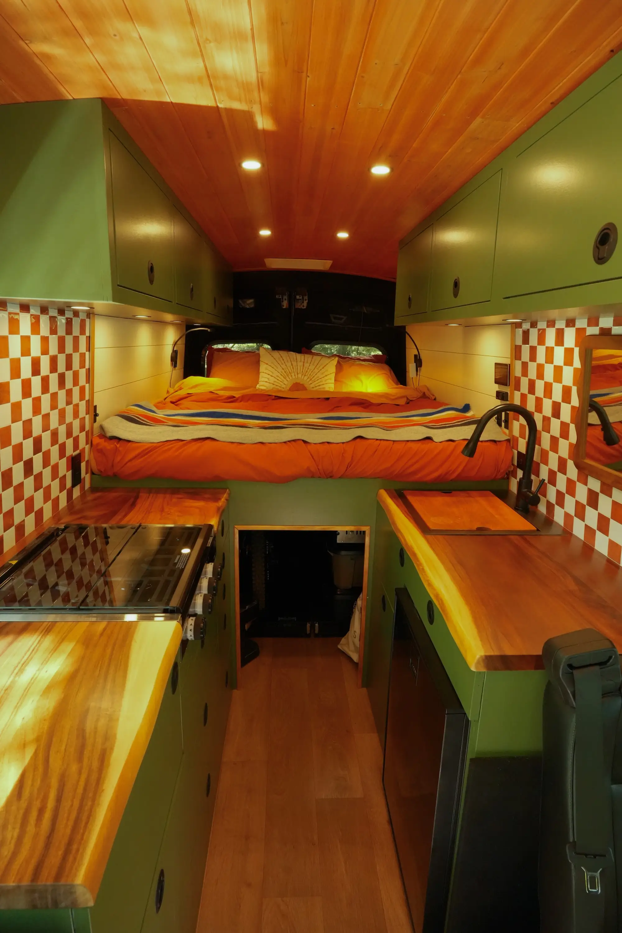 The interior of a van that has been turned into a living space. It has a large bed, green cabinetry, a wooden ceiling, wooden countertops, and an orange checkerboard backsplash.