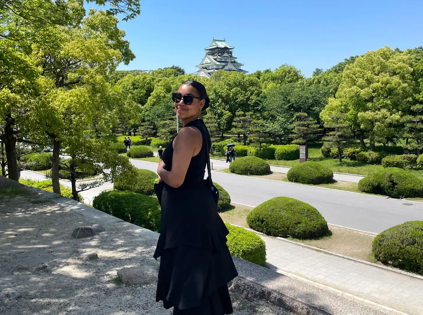 The writer wearing a black dress and sunglasses and standing in front of green bushes in Osaka, Japan.