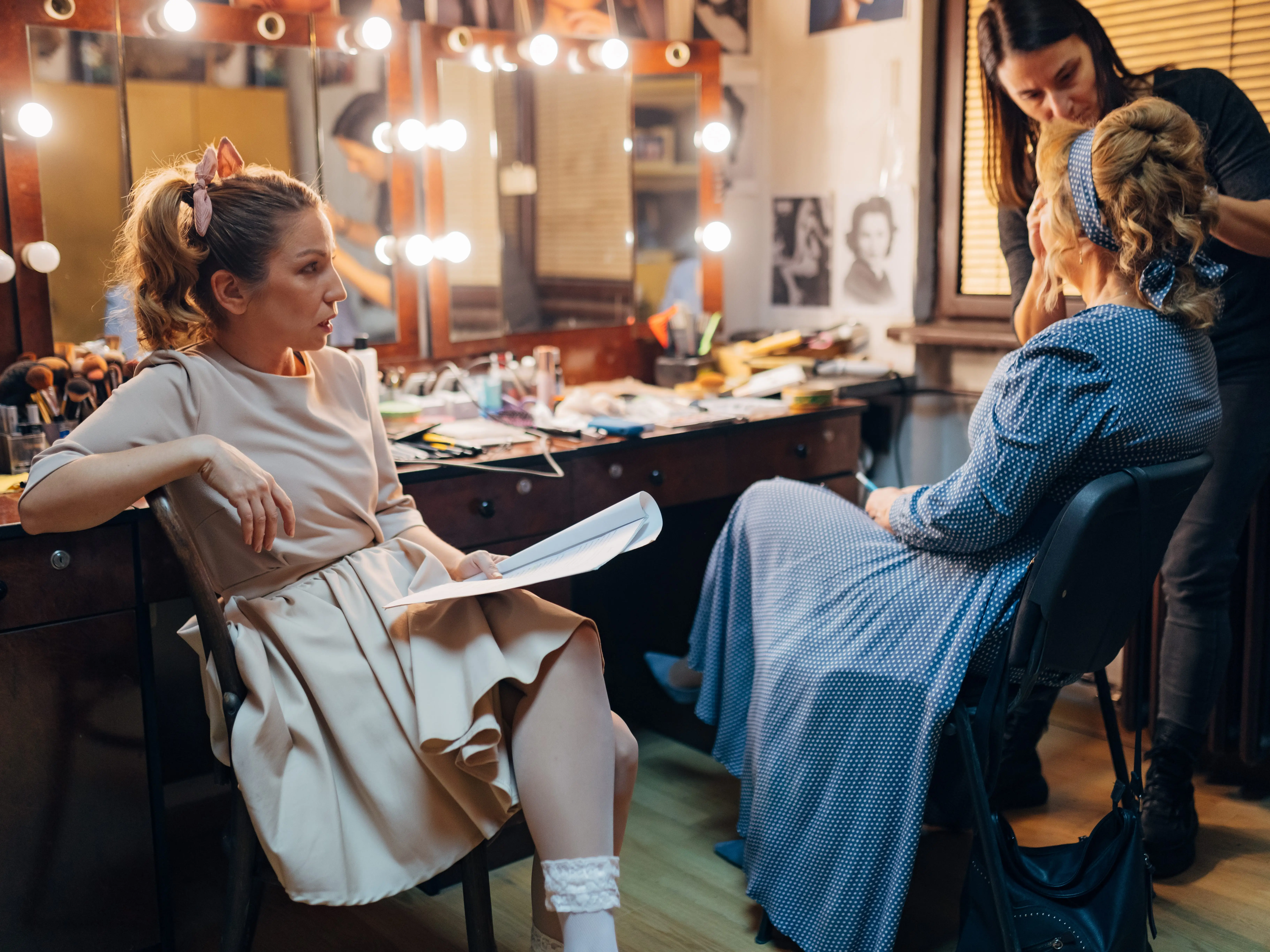 A makeup artist applies makeup to someone who is wearing a blue dress while another person sits nearby