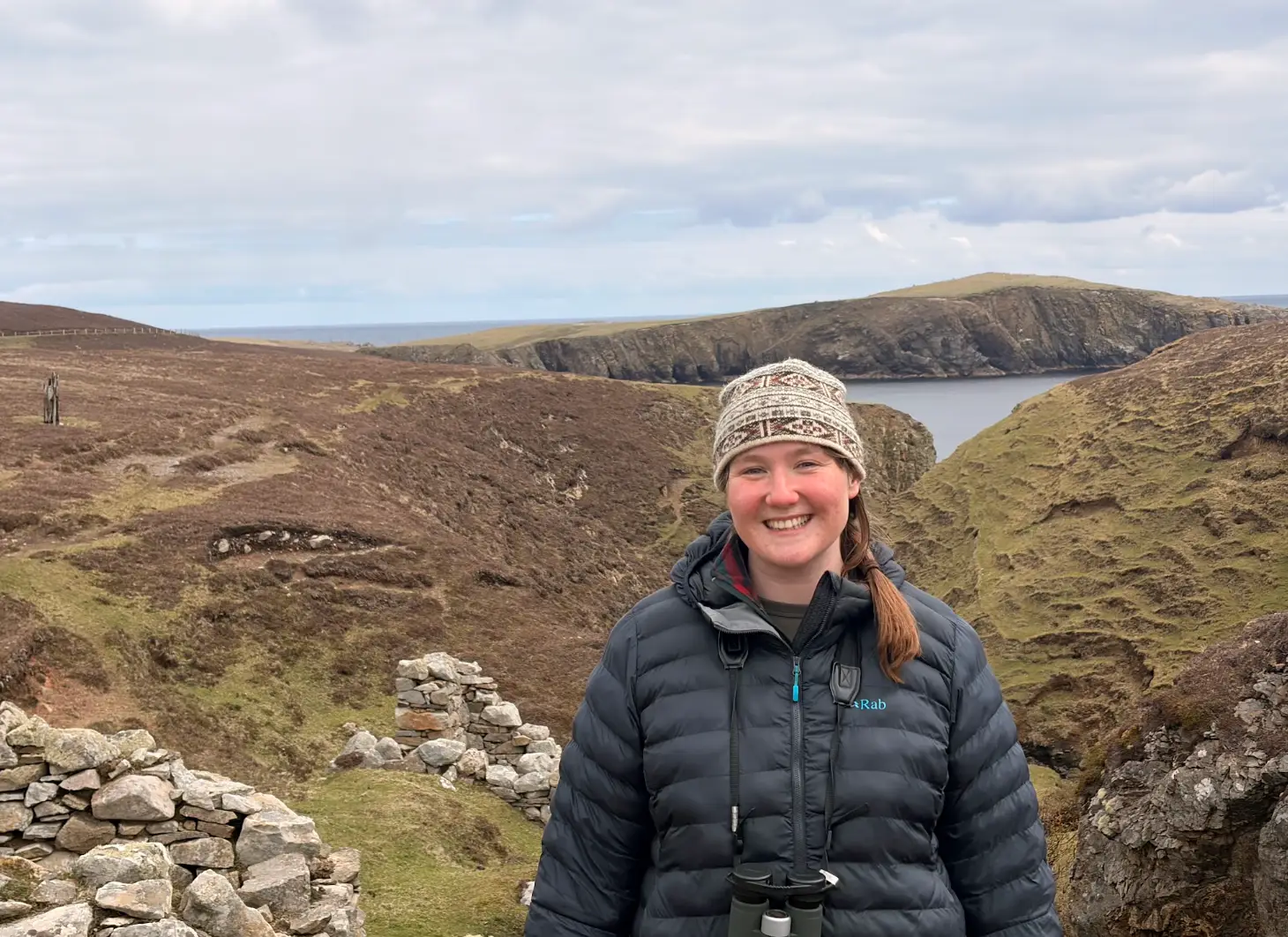 The writer wearing a coat and hat and standing in front of hills in Fair Isle.