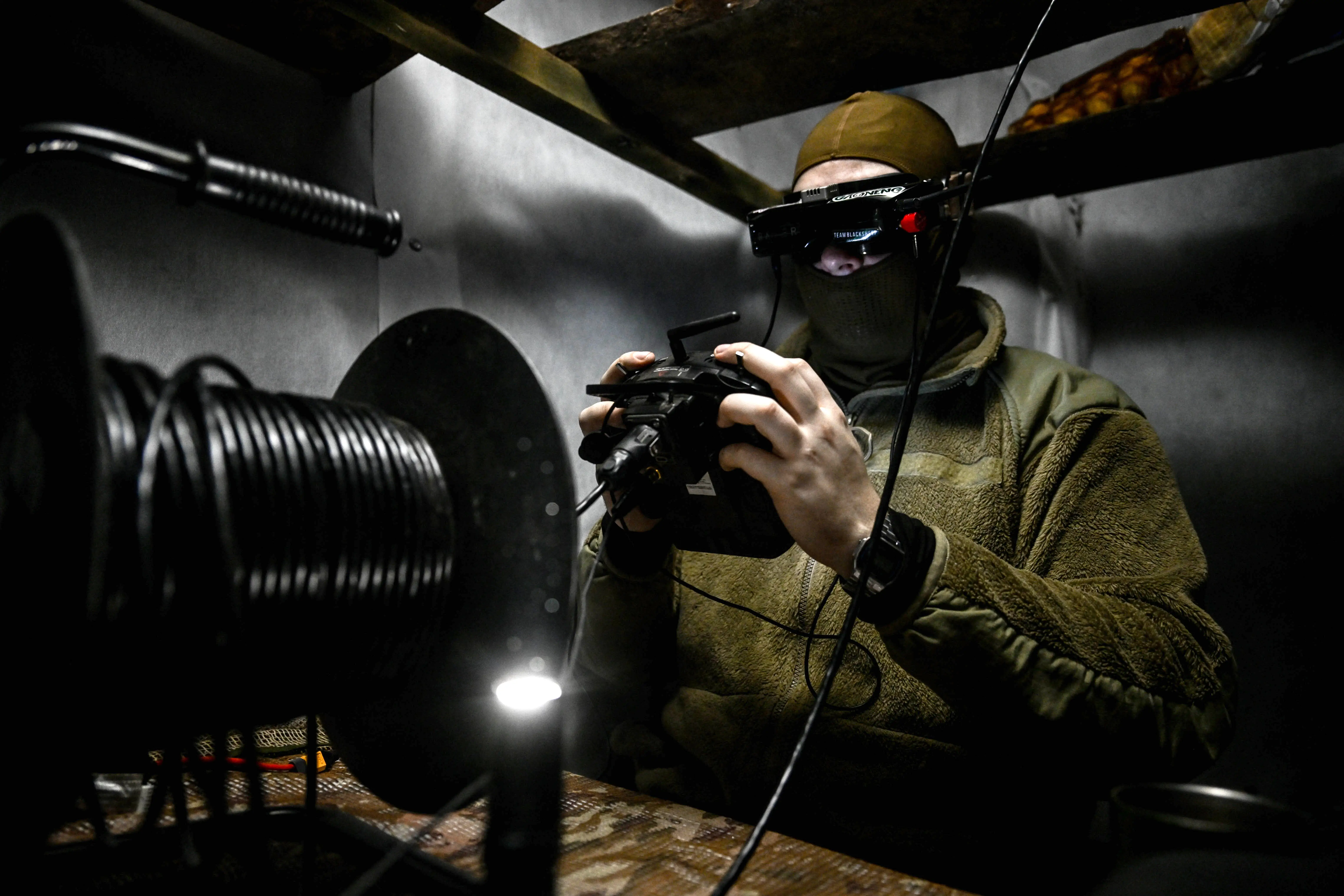 A man in khaki and a headset holds a controller in a small grey enclosed space with a black spool of cable in front of him