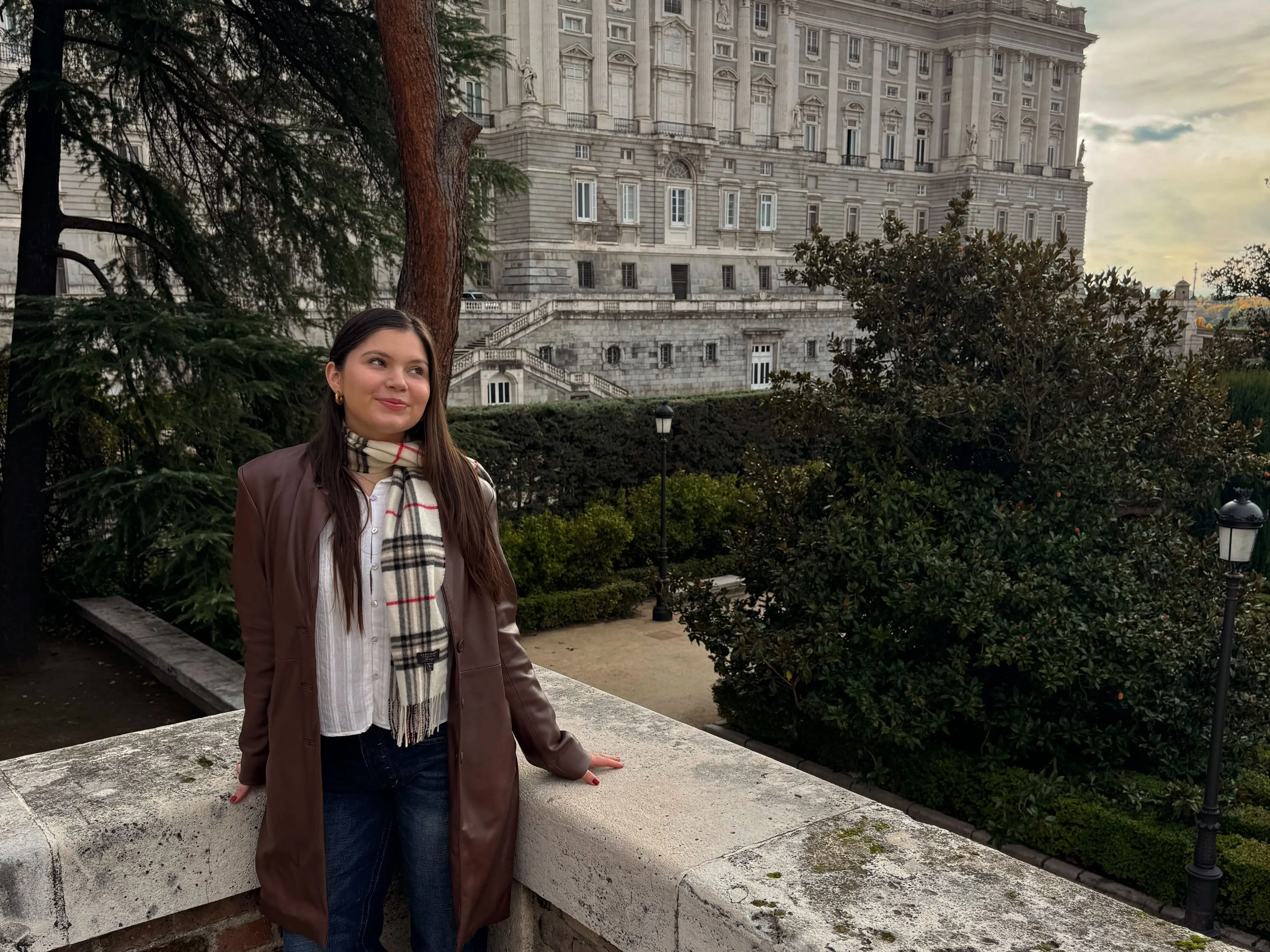 The writer wearing a brown coat and standing in front of trees and a building in Madrid, Spain.