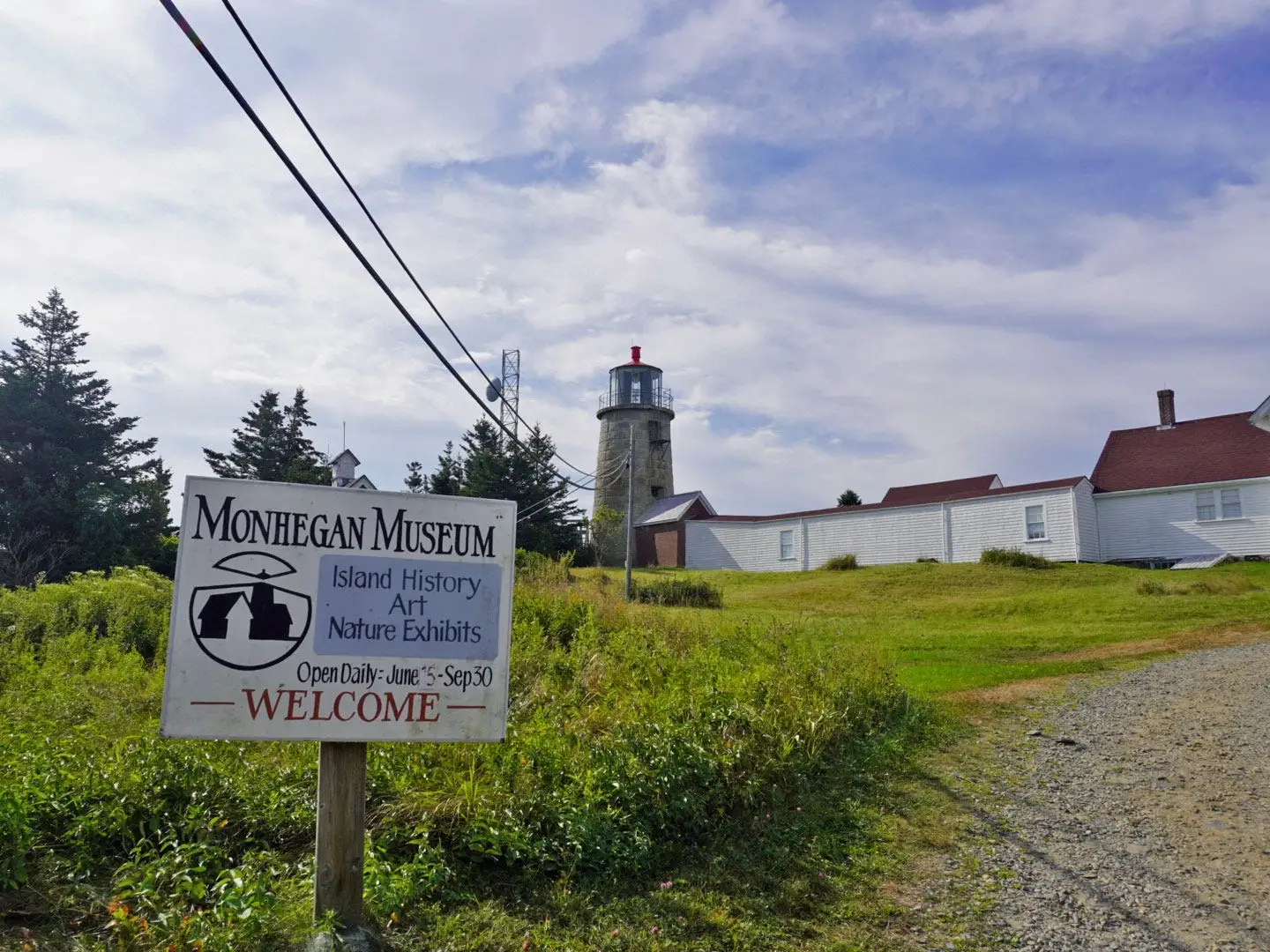 A photo of the Monhegan Museum welcome sign, with a lighthouse in the background.