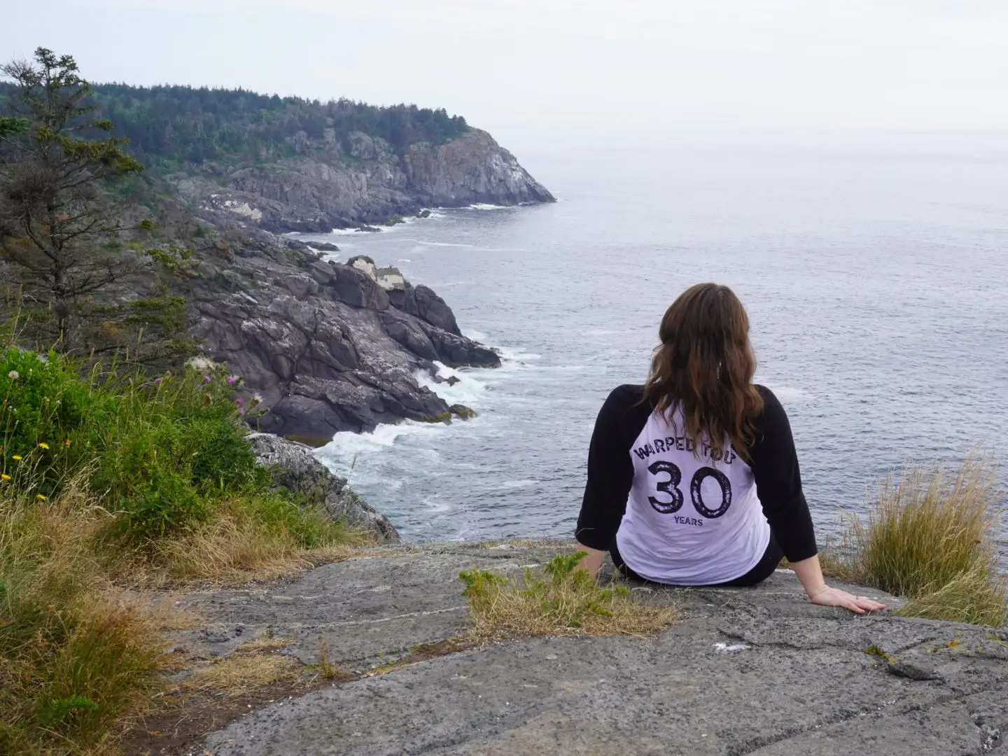 Emy sits on a rock, looking out at the ocean and cliffs.
