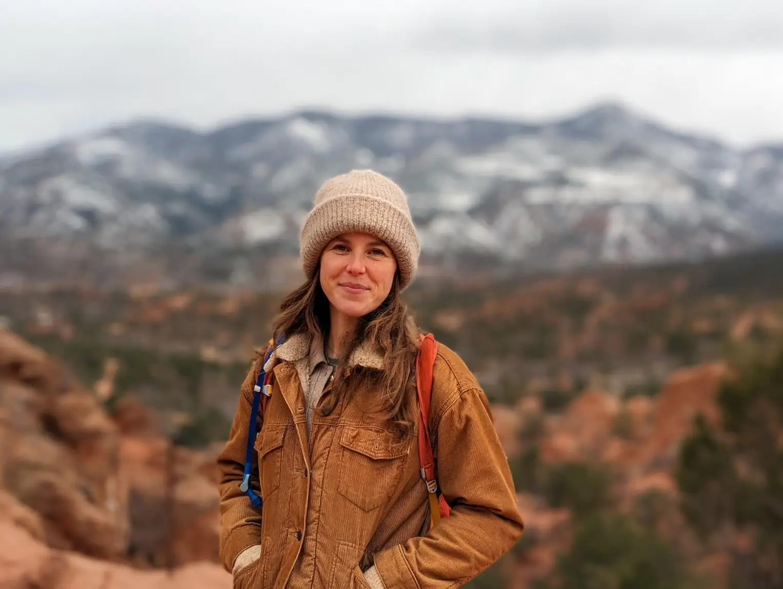 The writer wearing a hat and brown jacket and standing in front of a mountain.