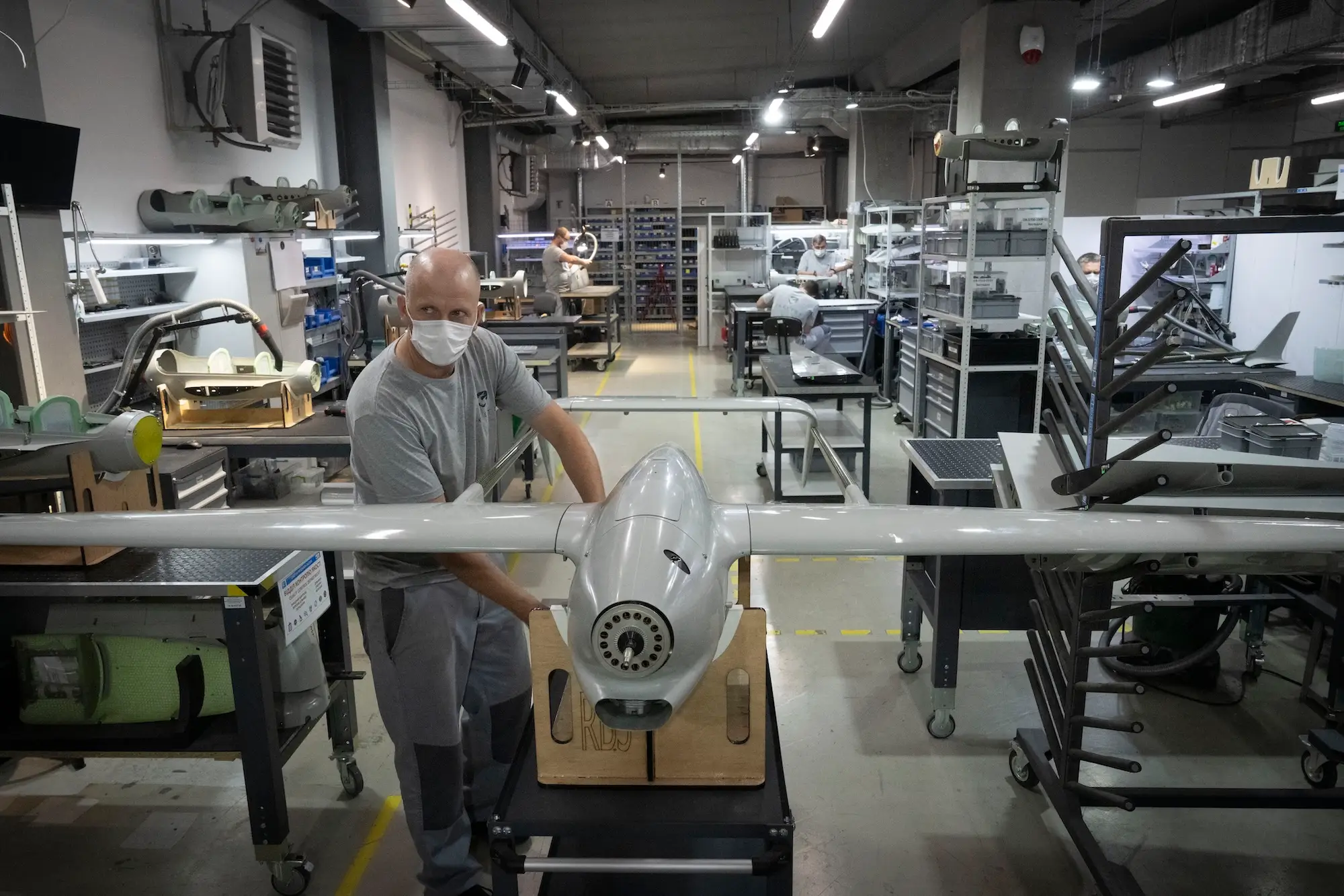 A man in a grey t-shirt and trousers and a white mask stands beside a large grey drone inside a grey room with drone parts and shelving