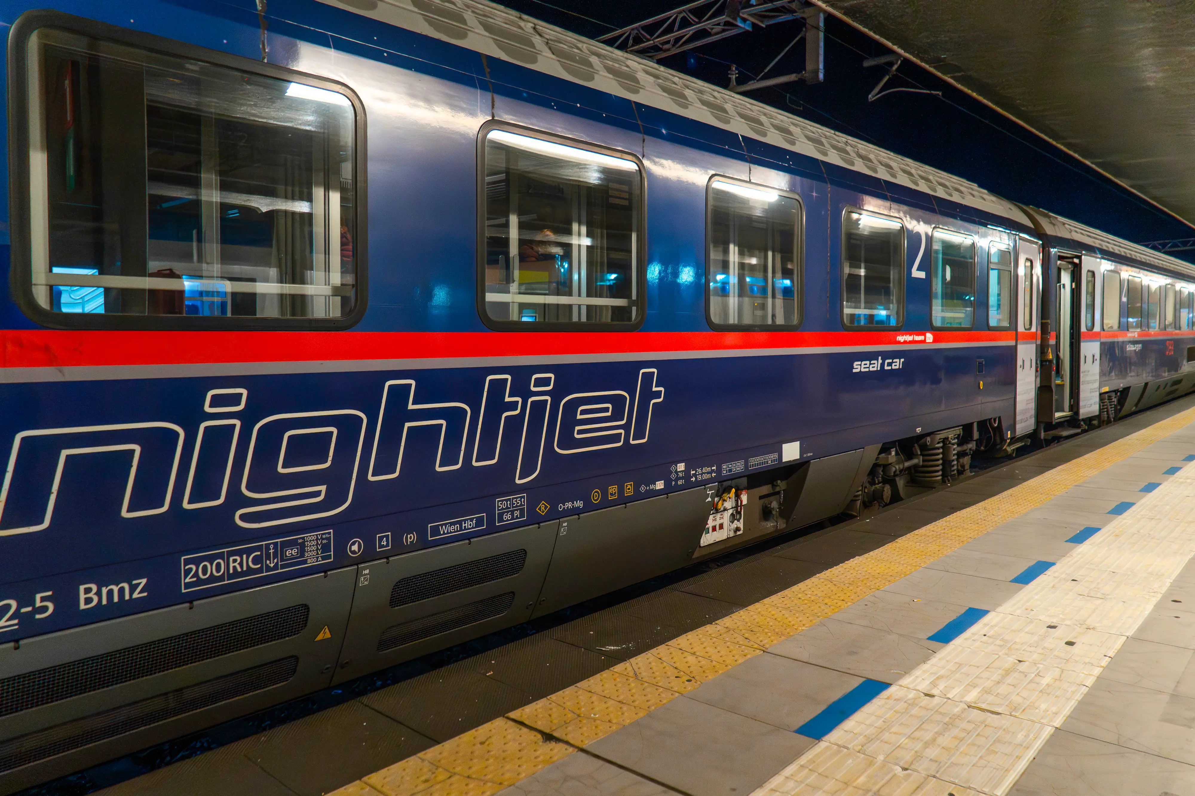 A navy blue Nightjet train parked at a lit-up outdoor platform at night