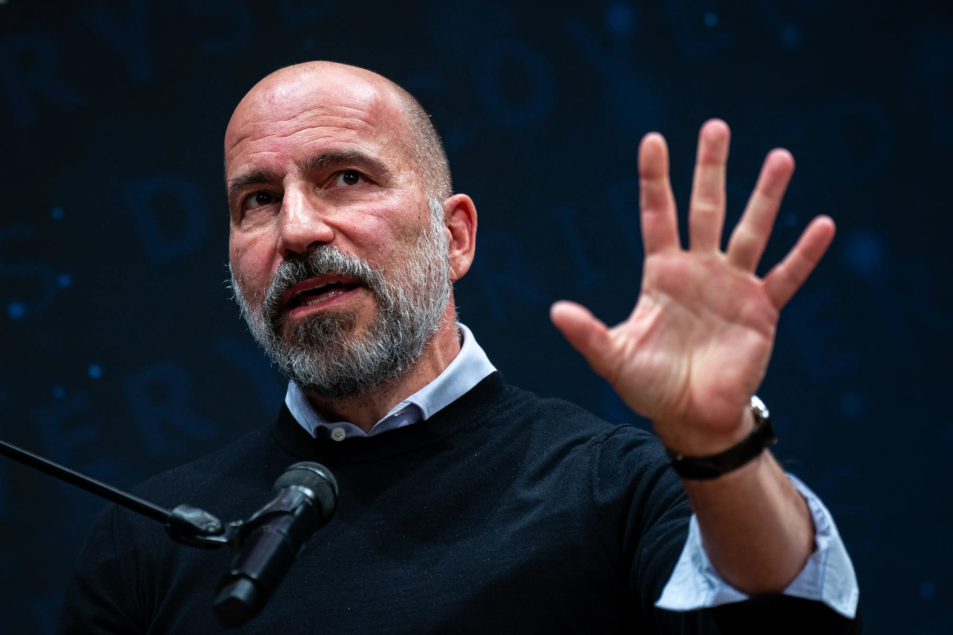 Dara Khosrowshahi speaks at a microphone while raising his left hand and wearing a light blue dress shirt under a dark blue sweater.