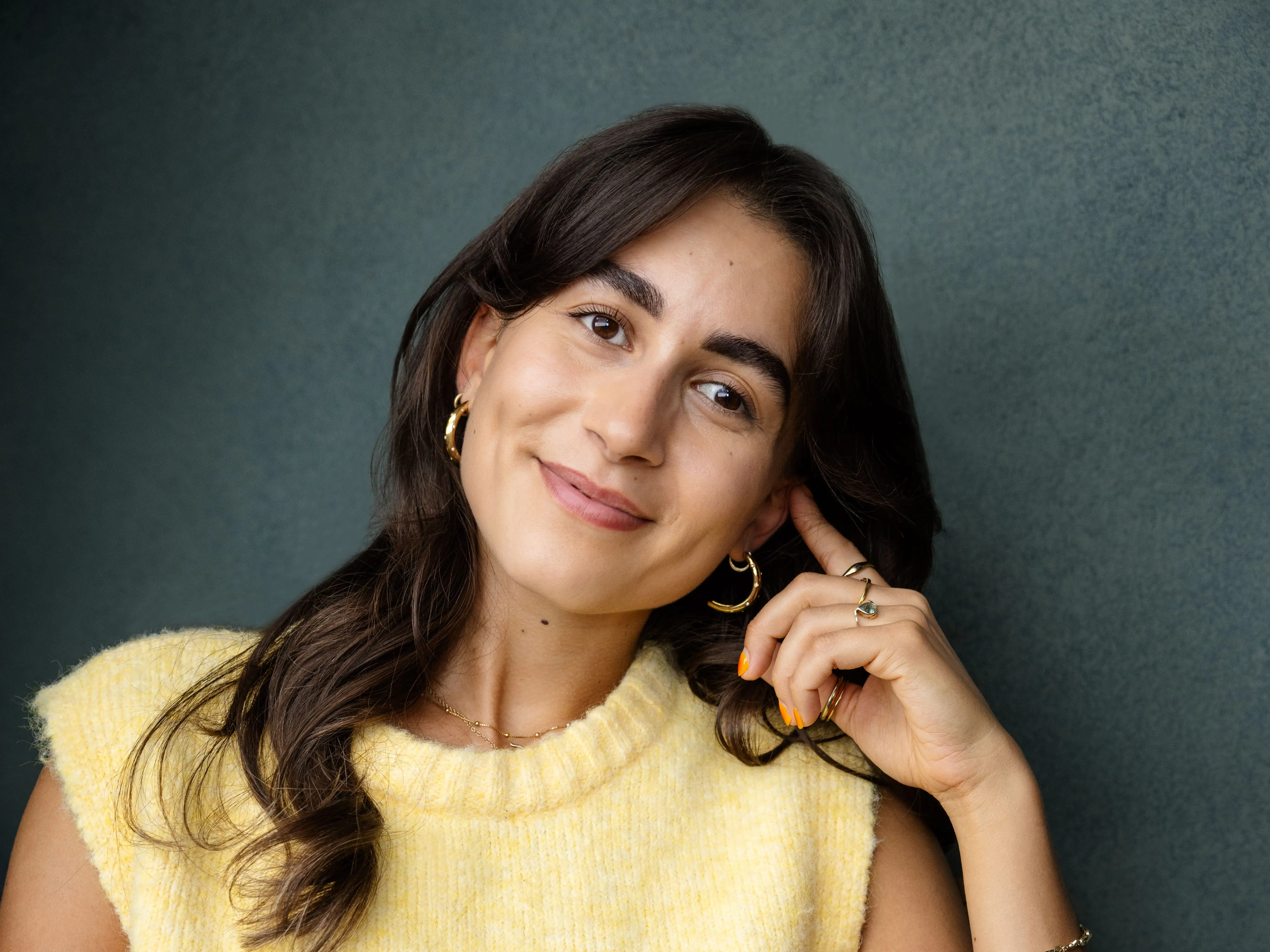 A woman wears a yellow sweater vest and sits in a wicker chair.