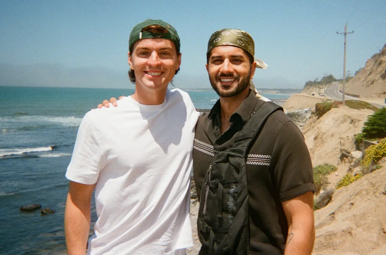 Two people smile with their arms around each other on a coastal cliffside overlooking the ocean and a winding shoreline road.