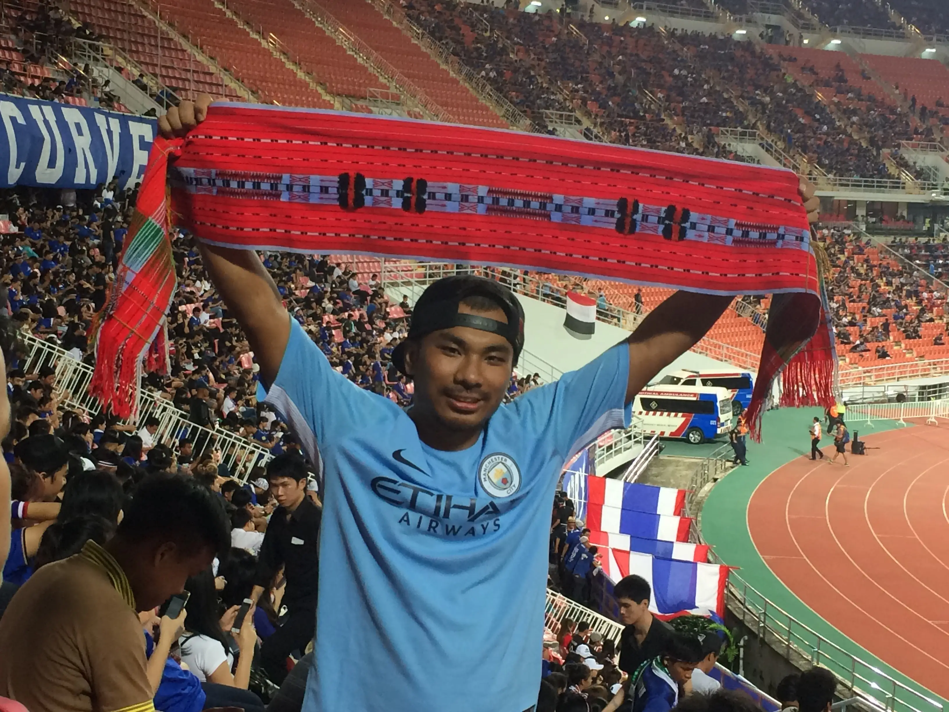 Worchihan Zingkhai in a stadium wearing a blue shirt and holding up a red scarf.