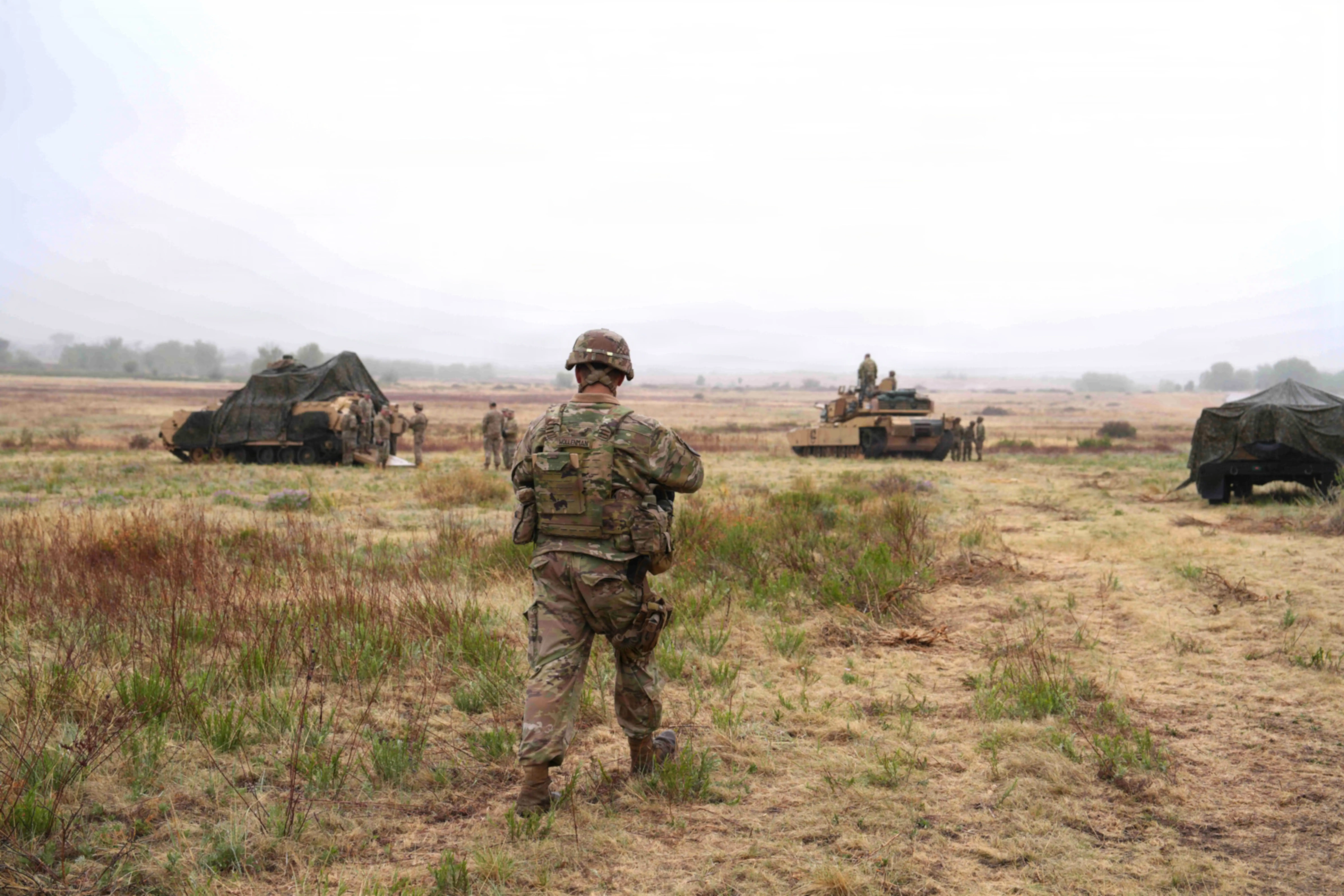 A soldier walks on green and brown grass with tanks and vehicles in front of him and a cloudy sky.