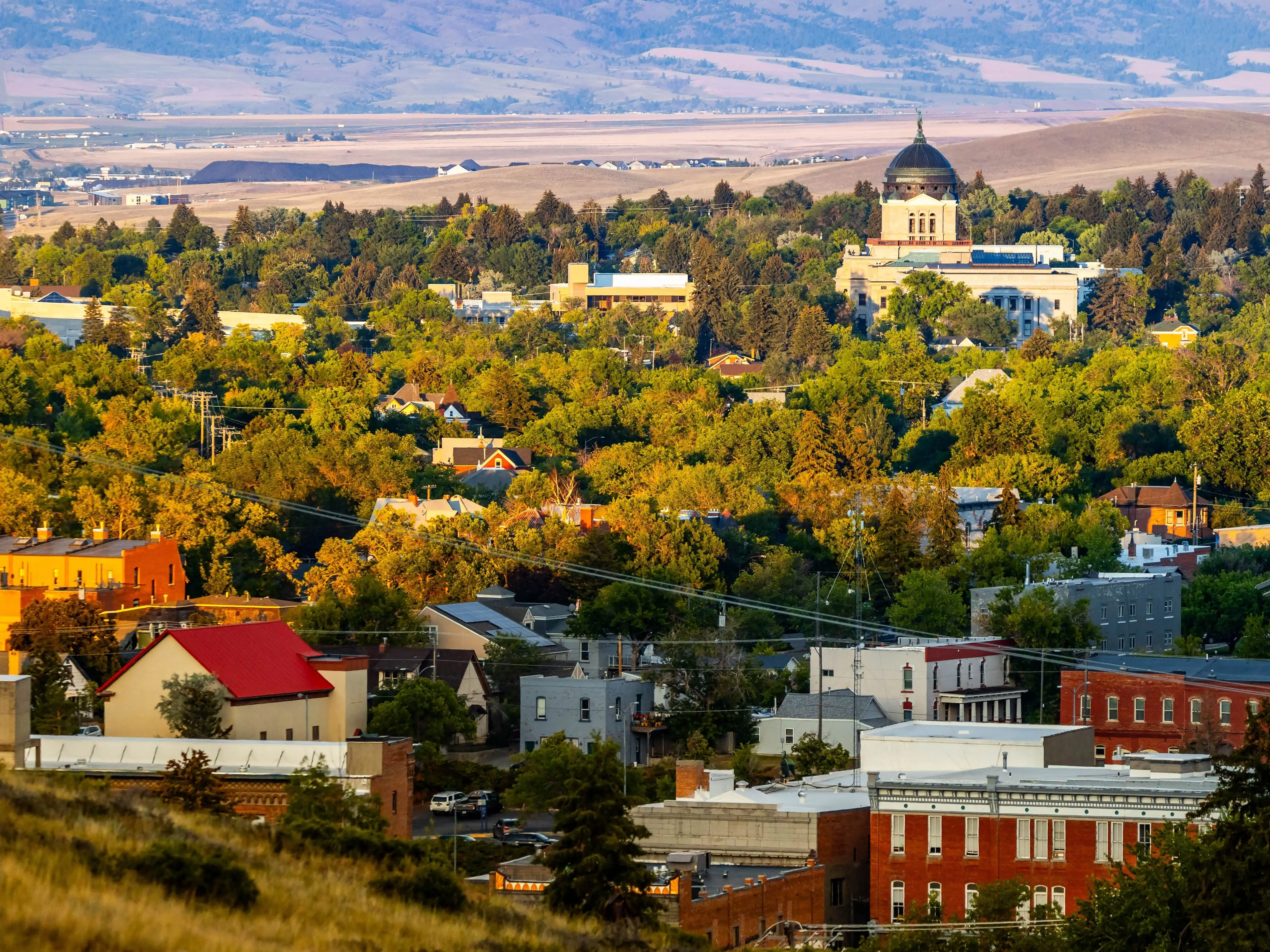 A panoramic aerial view of the city of Helena, Montana, is captured at sunset, showcasing the urban landscape and the iconic State Capitol under the warm evening light