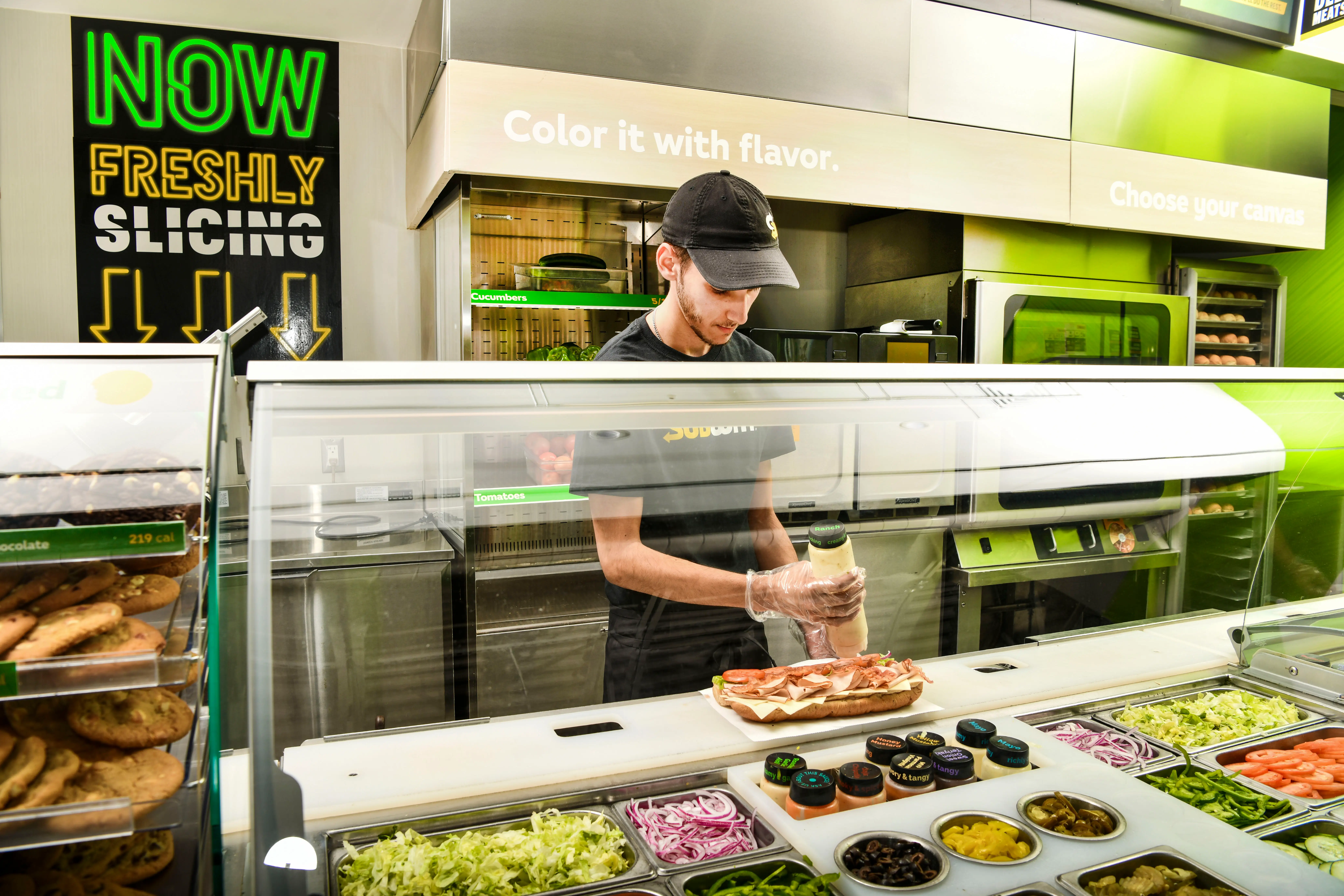 A worker prepares a sandwich behind a counter at a Subway restaurant while wearing a black baseball cap, t-shirt, and pants. In front of him sit containers with ingredients such as sliced tomatoes, red onions, shredded lettuce, and mayo.