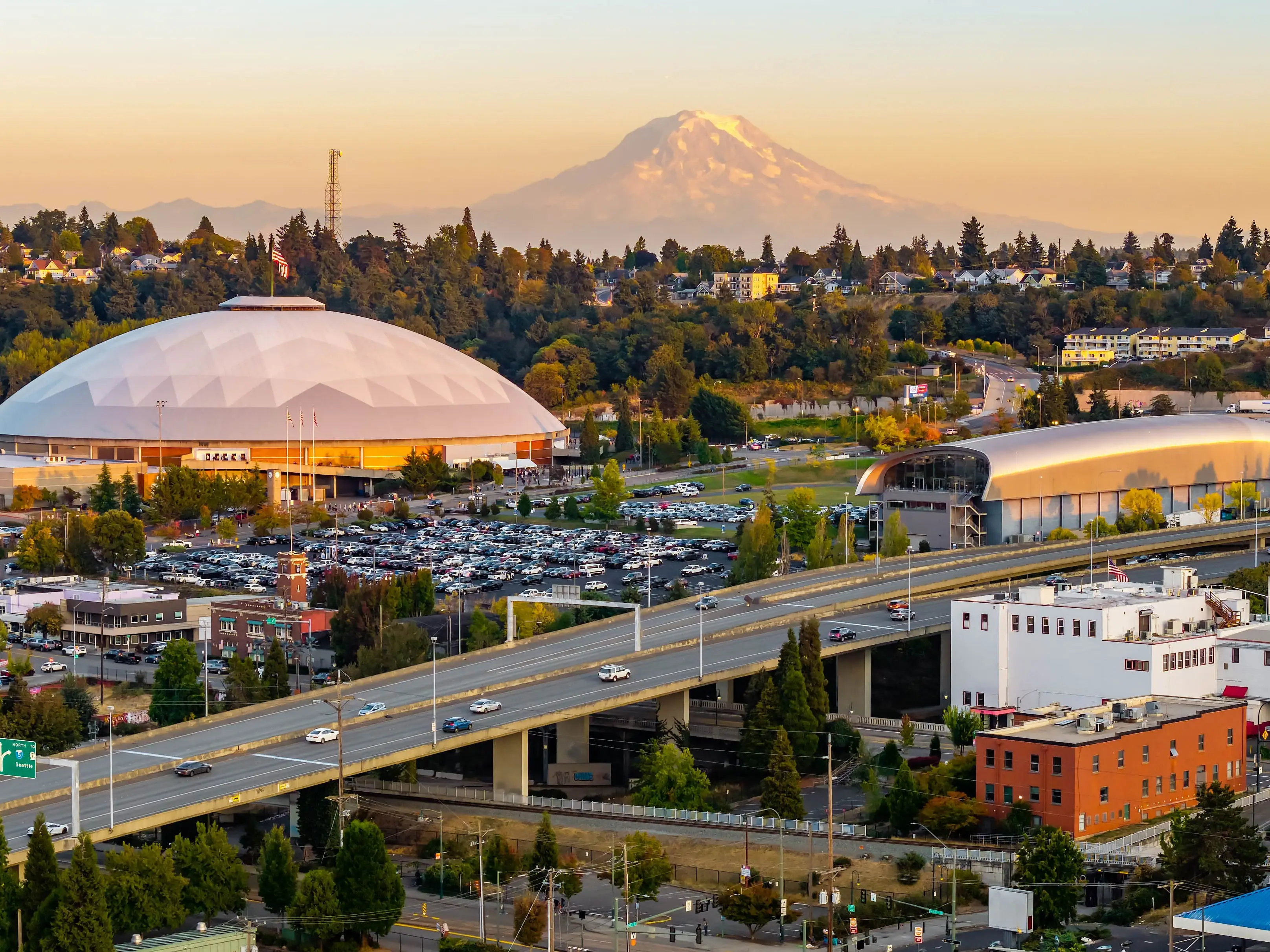 The Tacoma cityscape and its skyscrapers are dramatically framed against the backdrop of the towering, snow-capped Mount Rainier at sunset.