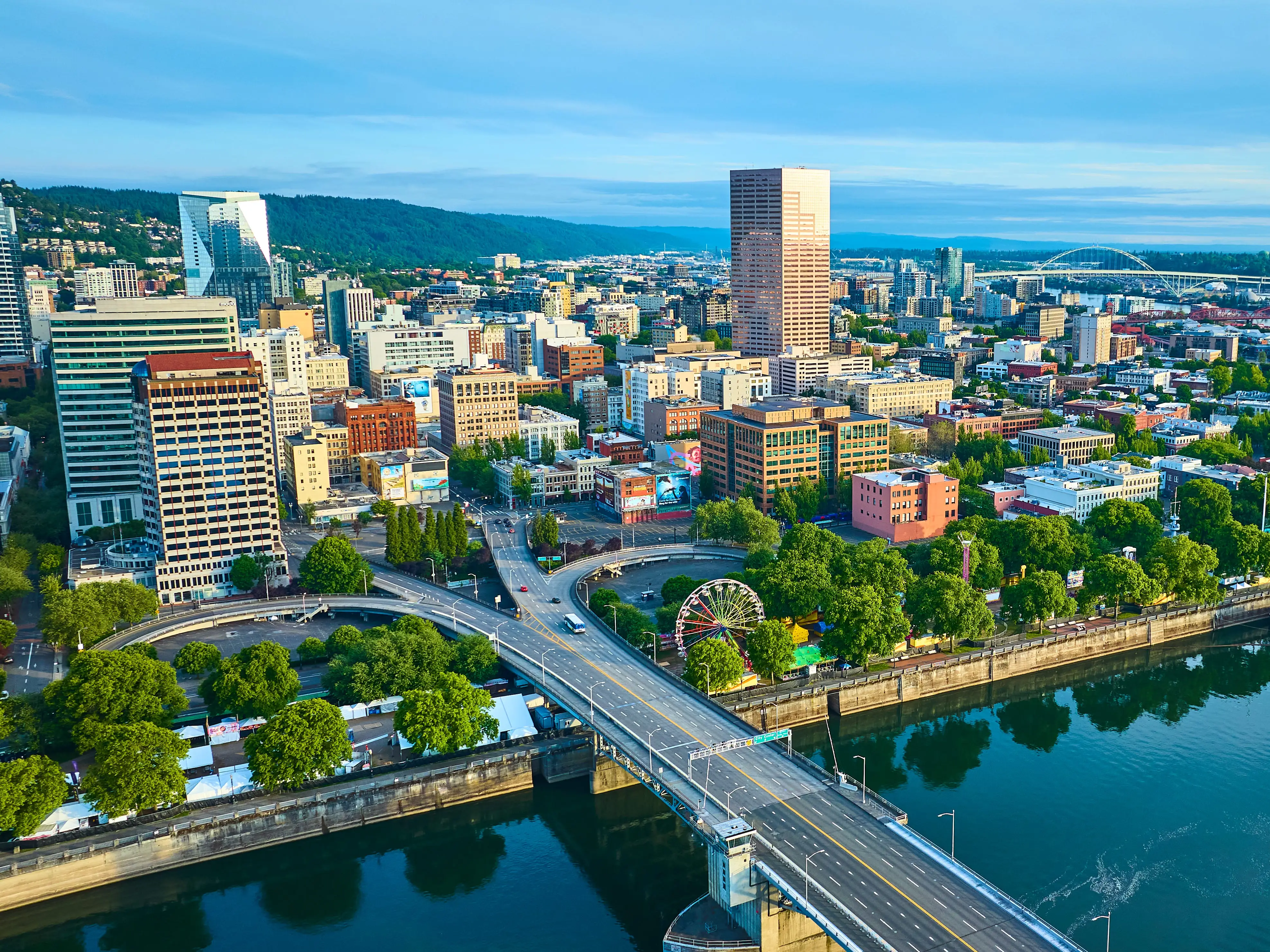 Aerial view of Portland, Oregon, showcasing the vibrant downtown with a mix of modern and classic skyscrapers. The Willamette River with a bridge and paddlewheel riverboat adds scenic beauty and historical charm.