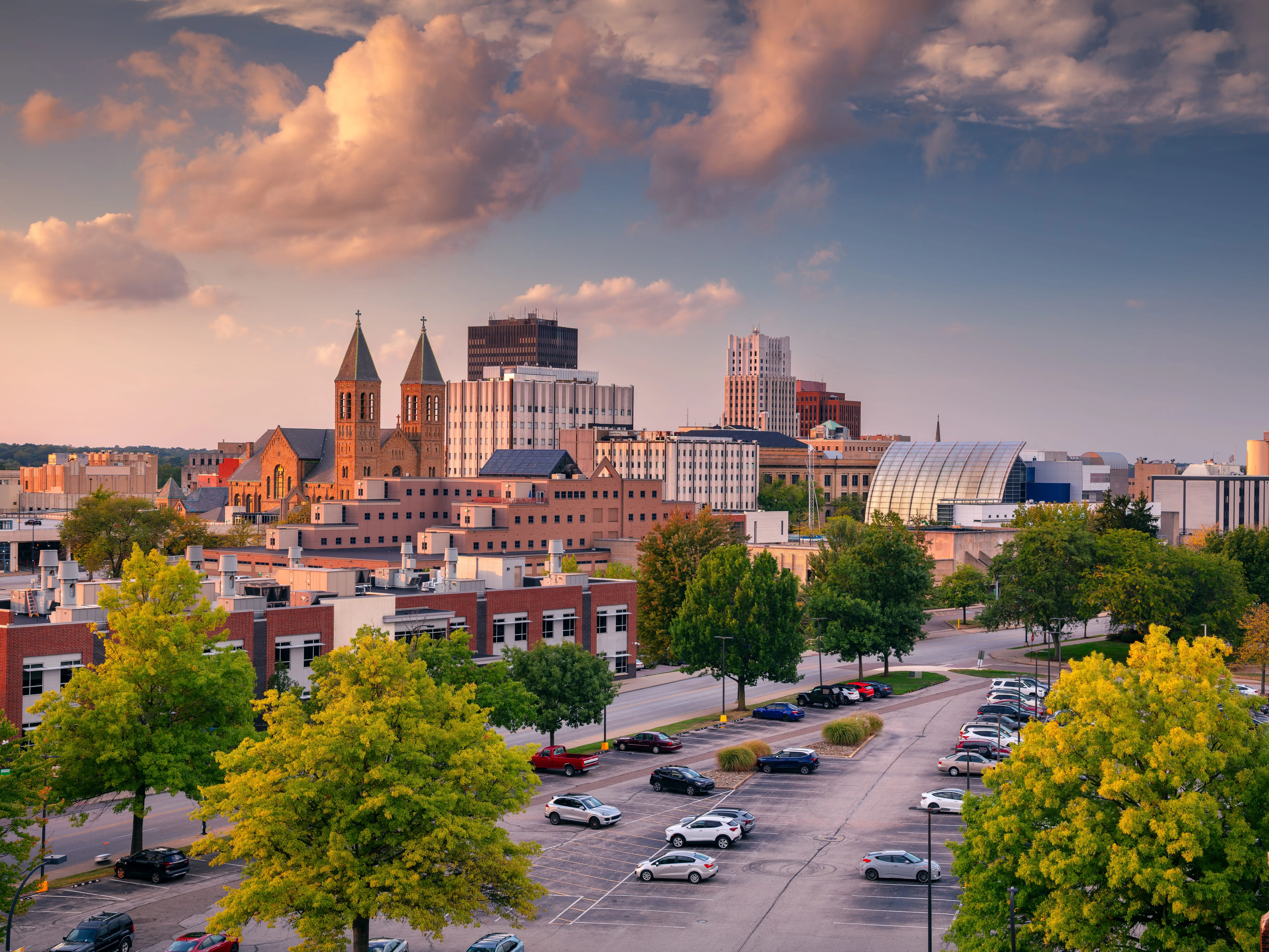 Downtown Akron, Ohio, USA at autumn sunset features a mix of modern and historic architecture. Prominent are twin-spired churches and contemporary buildings. The foreground shows a parking lot with evenly spaced cars and vibrant green trees. In the background, the skyline rises under a dramatic clouded sky, colored in warm hues by the setting sun. The scene captures a serene and picturesque moment in the city.