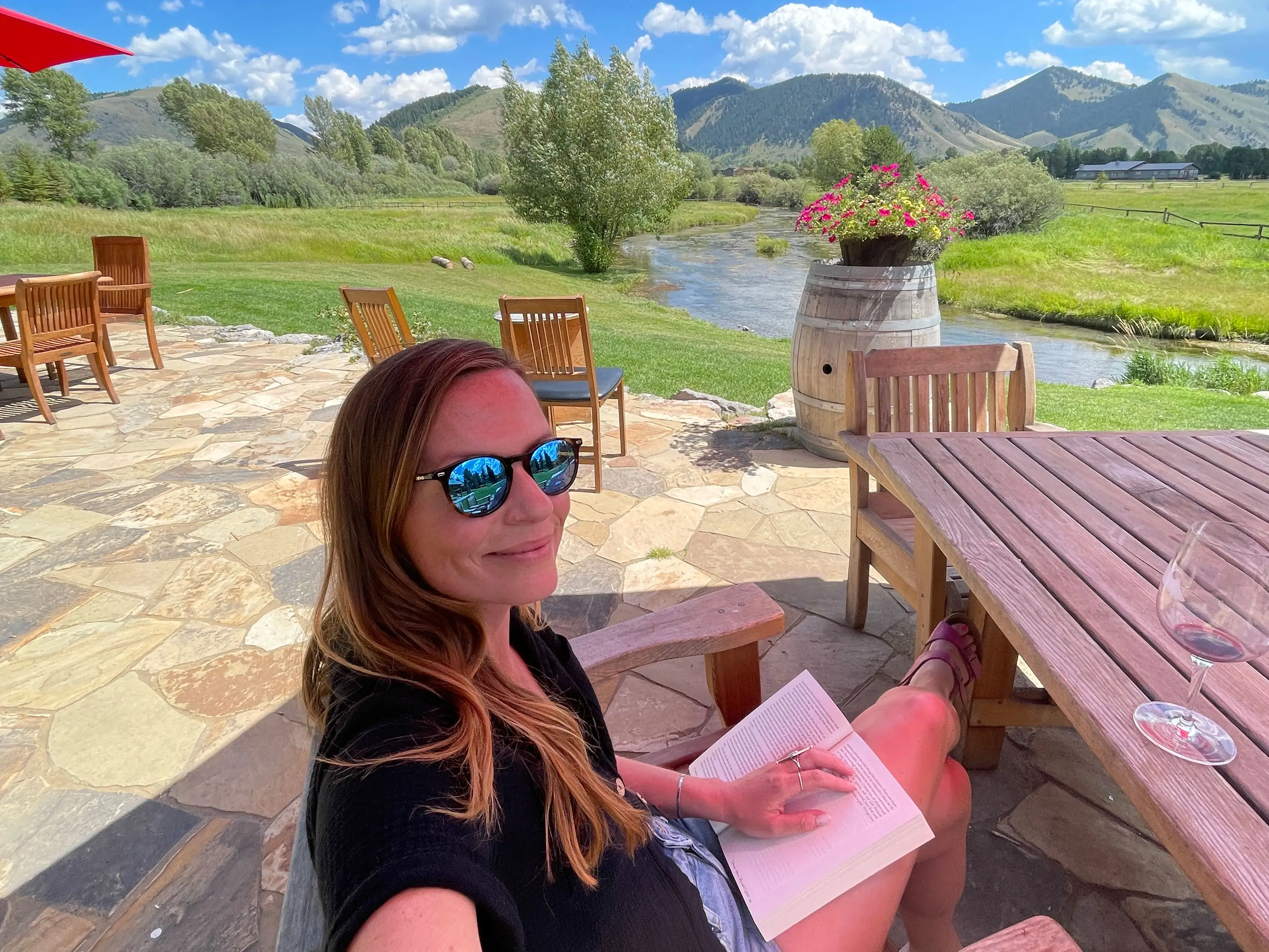 Emily reads a book on a patio with a river and mountains in the background.